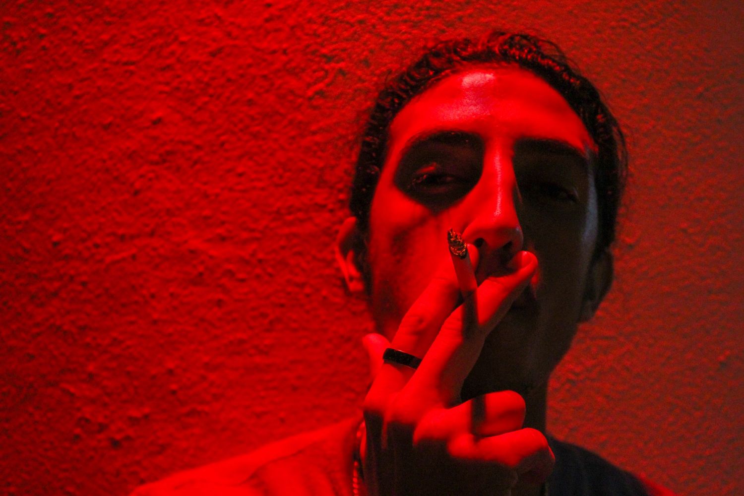 Close-up portrait of man smoking under intense red lighting against textured wall in dramatic low-key photography.