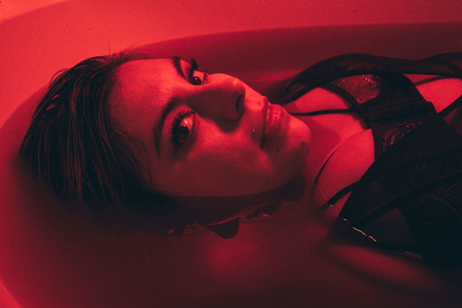 A woman with wet hair floats in a bathtub bathed in deep red light, wearing black lingerie.
