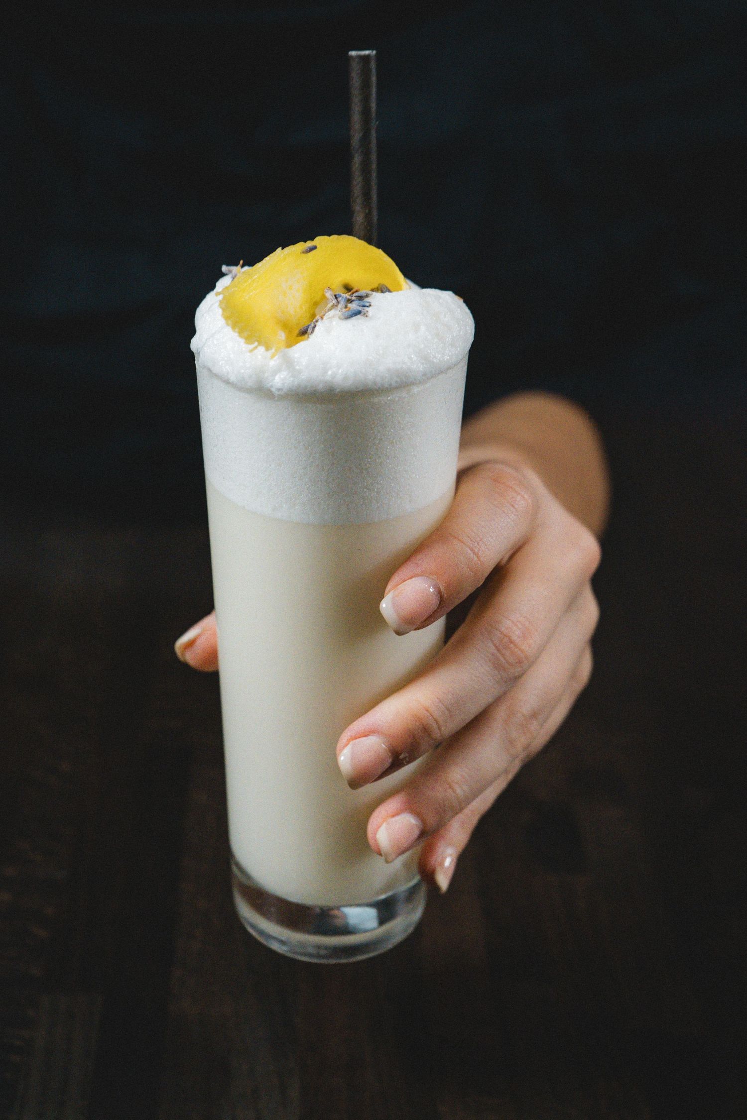 Hand holding tall frothy white cocktail with metal straw and yellow garnish on dark background.
