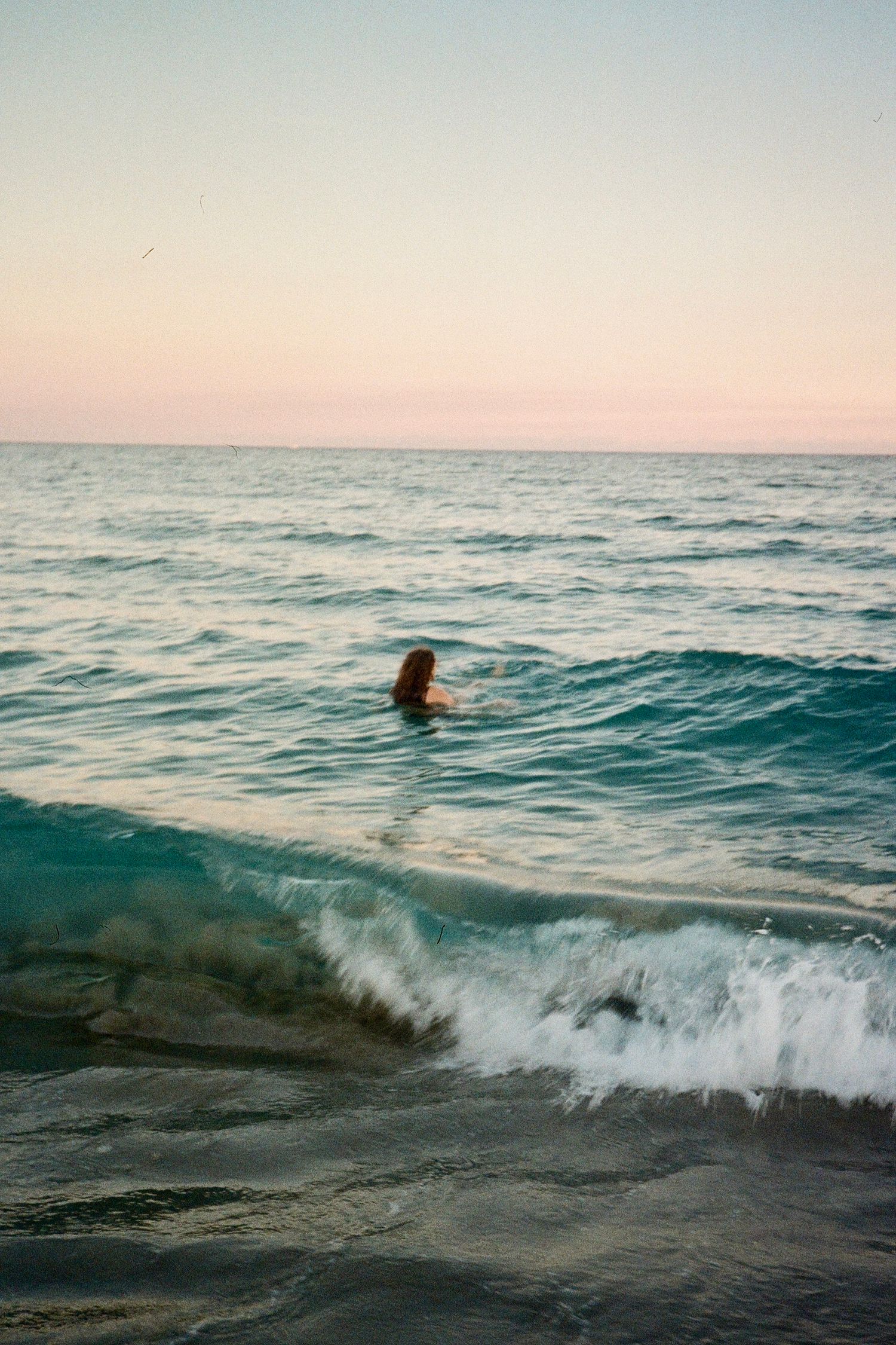 A person swims in turquoise ocean waves during a peaceful sunset at the beach.