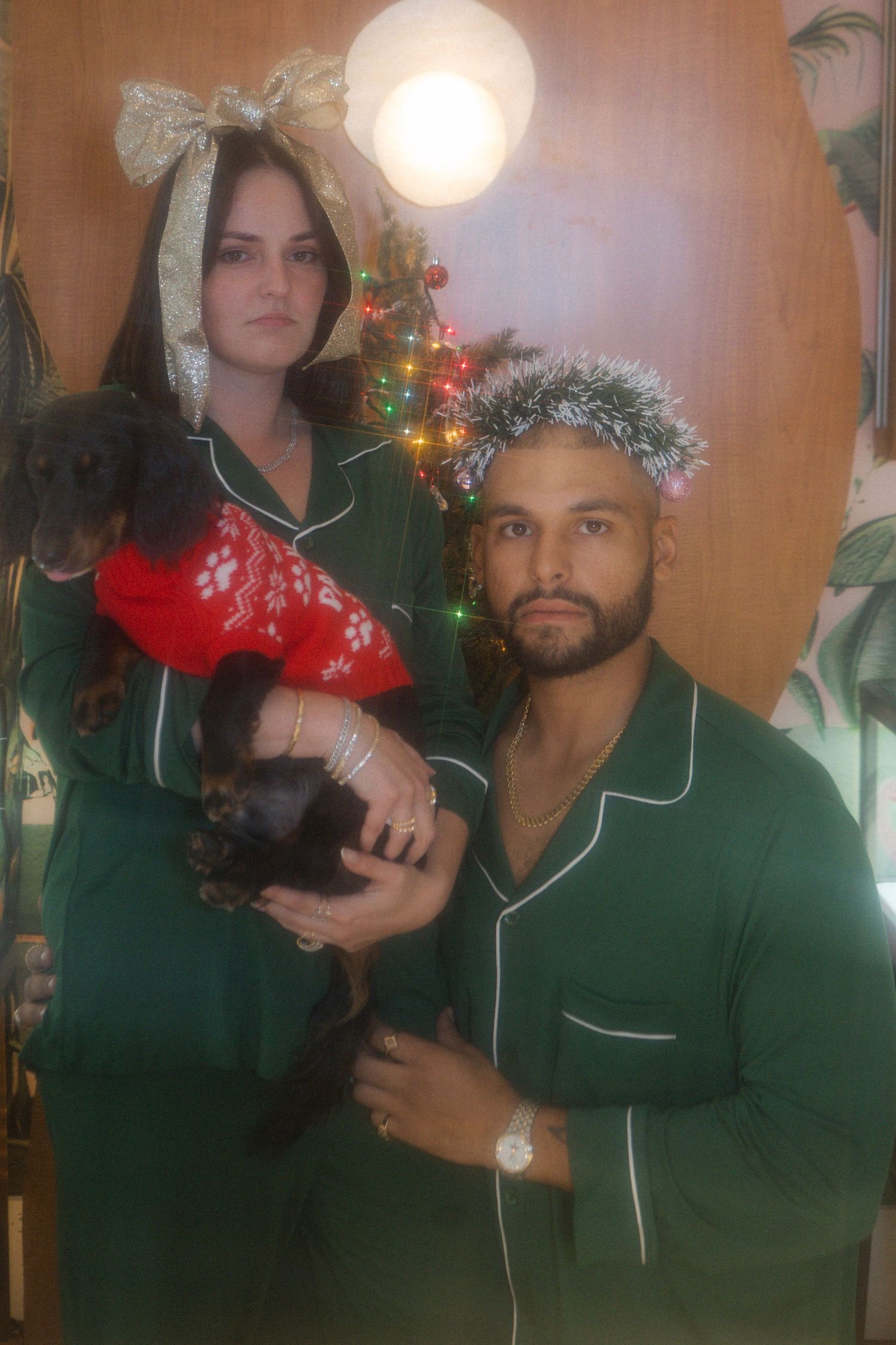 Couple in green Christmas pajamas holding a dachshund in red sweater near decorated holiday tree.