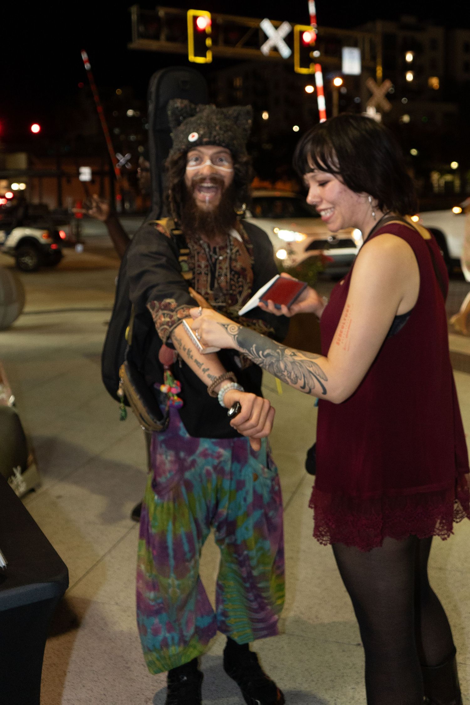 A tattooed man in tie-dye pants and pirate hat chats with a woman in a red dress on a city street at night.