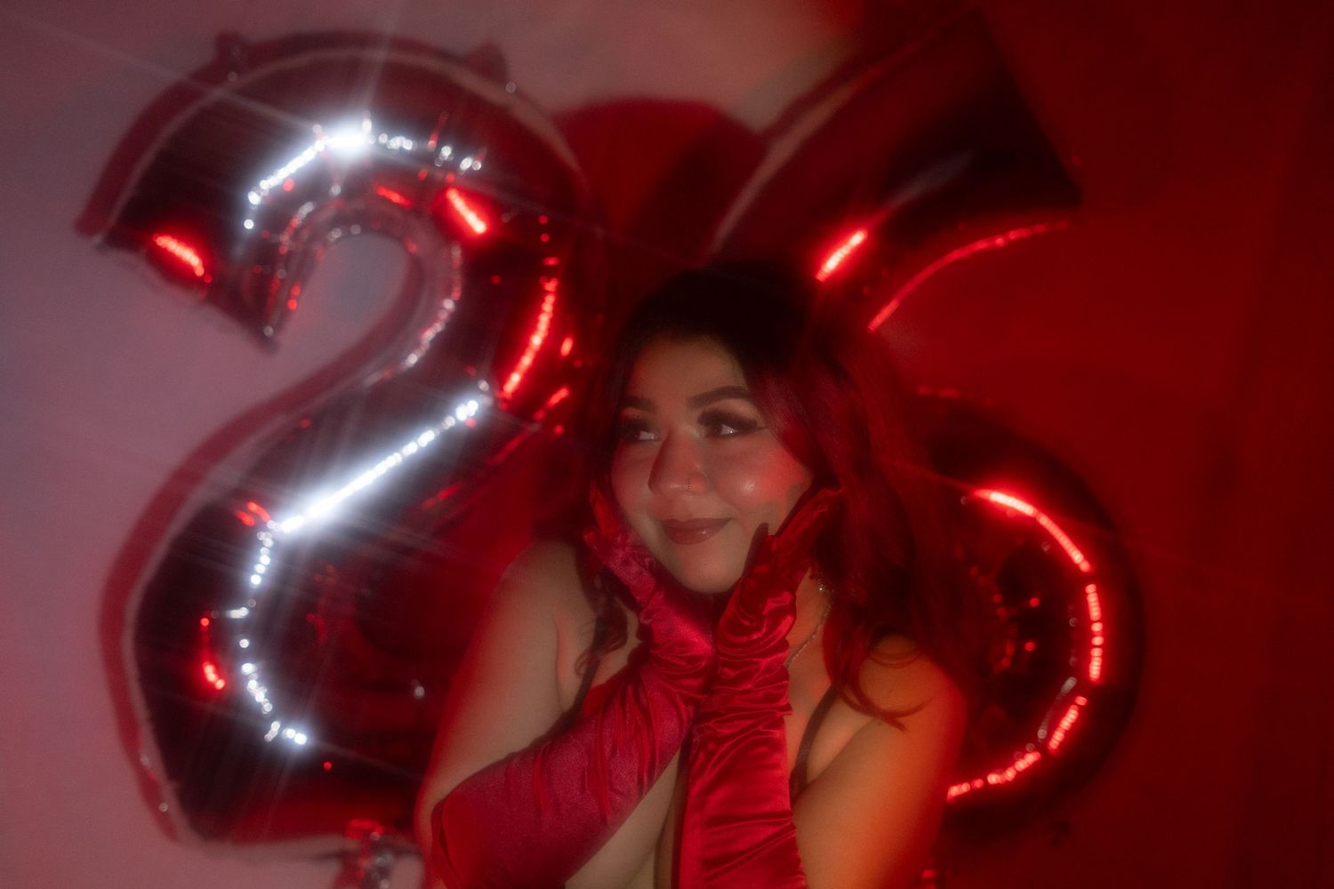 Woman with red hair lying next to silver number 26 balloons in a red-lit room.