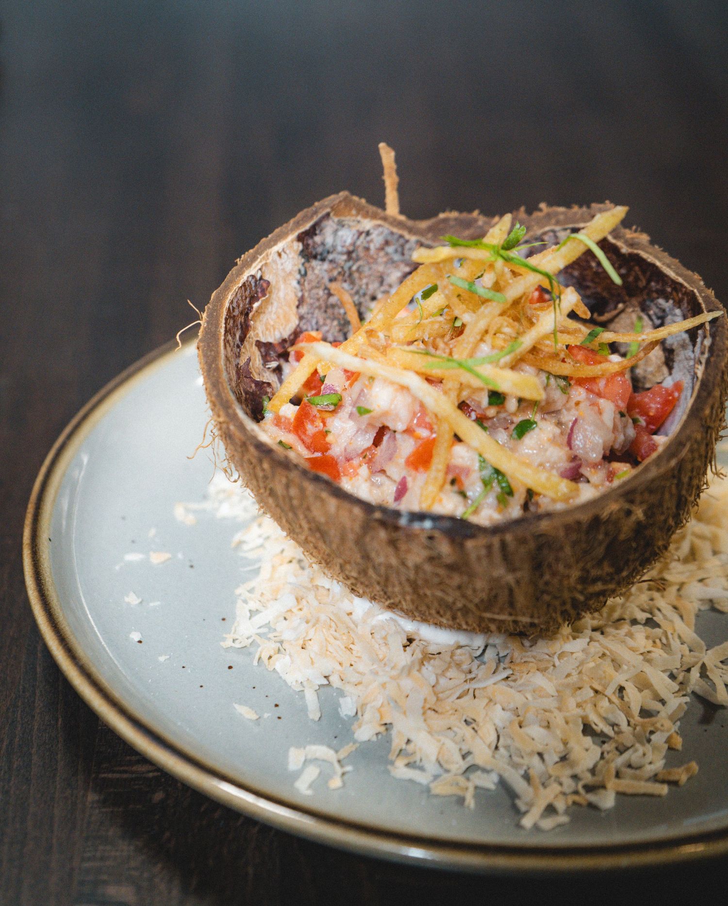 Gourmet seafood ceviche served in coconut shell bowl on bed of shredded coconut on ceramic plate.