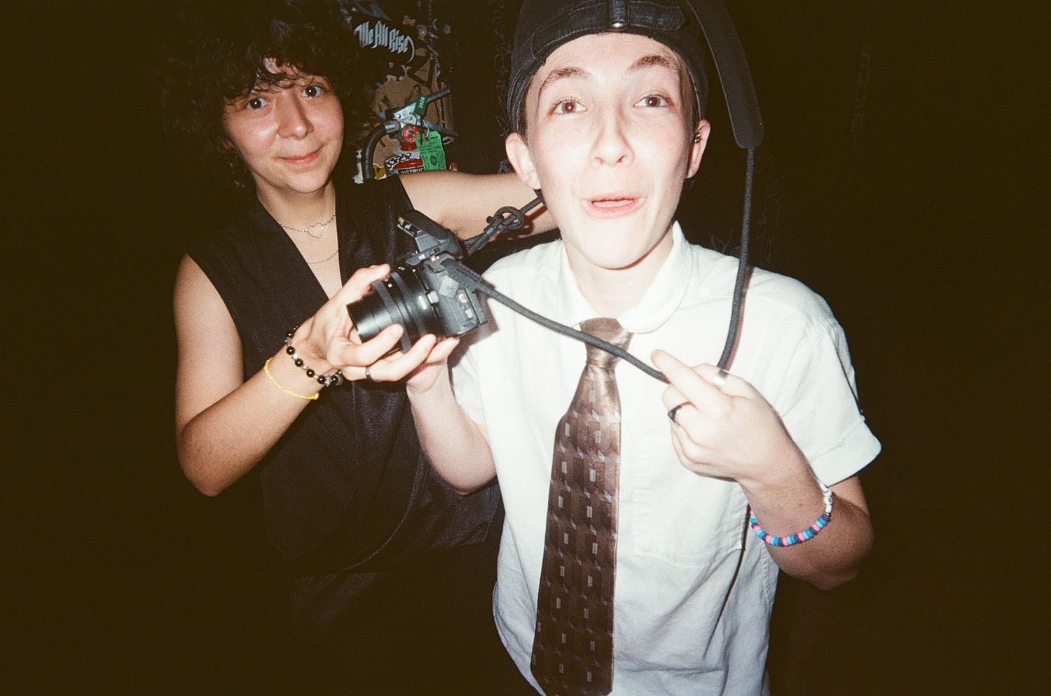 Two young people at a party, one holding a camera, the other wearing a tie and beanie hat indoors.