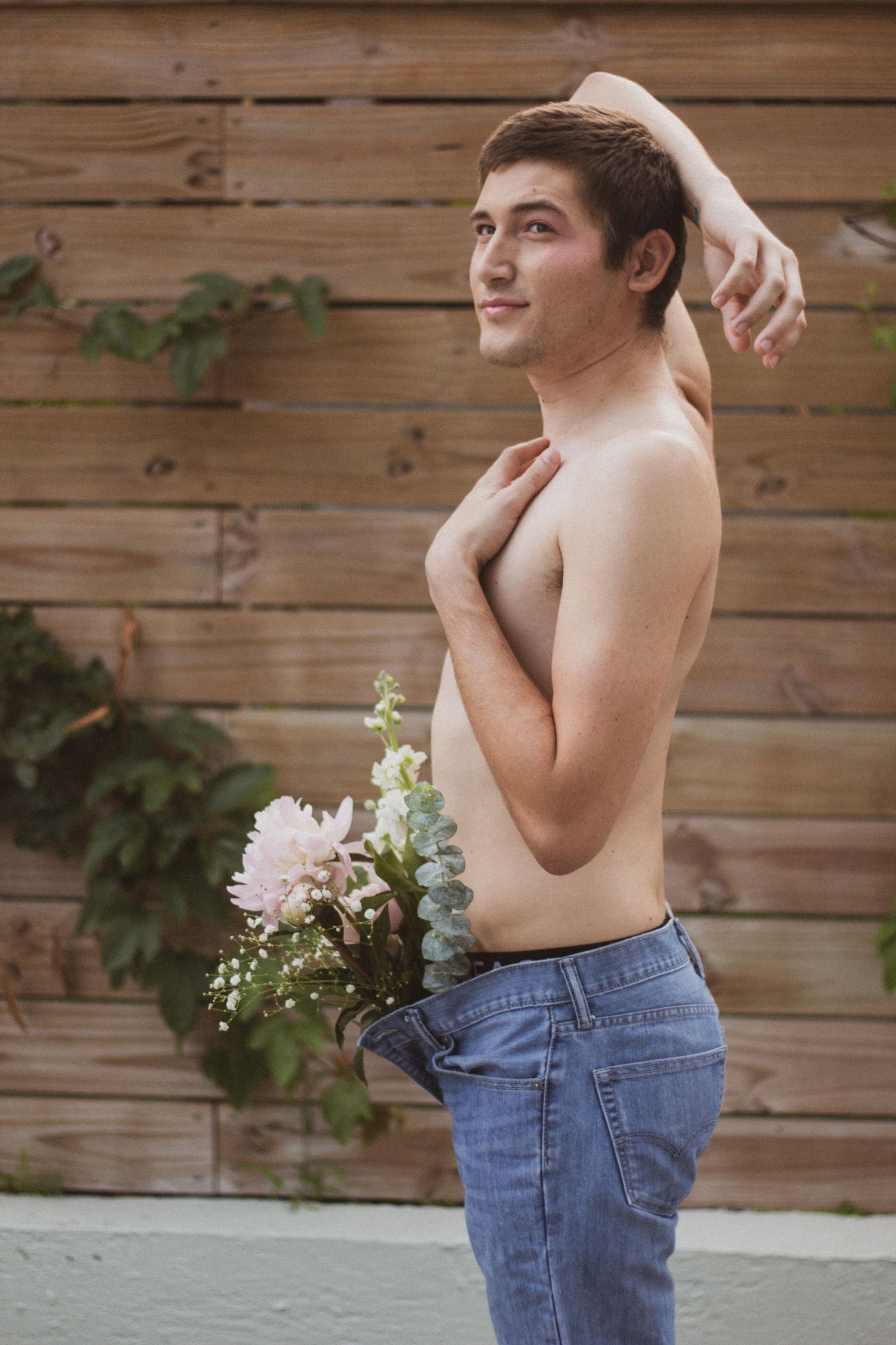 A shirtless young man poses with pink flowers tucked in his jeans beside a wooden fence with greenery.