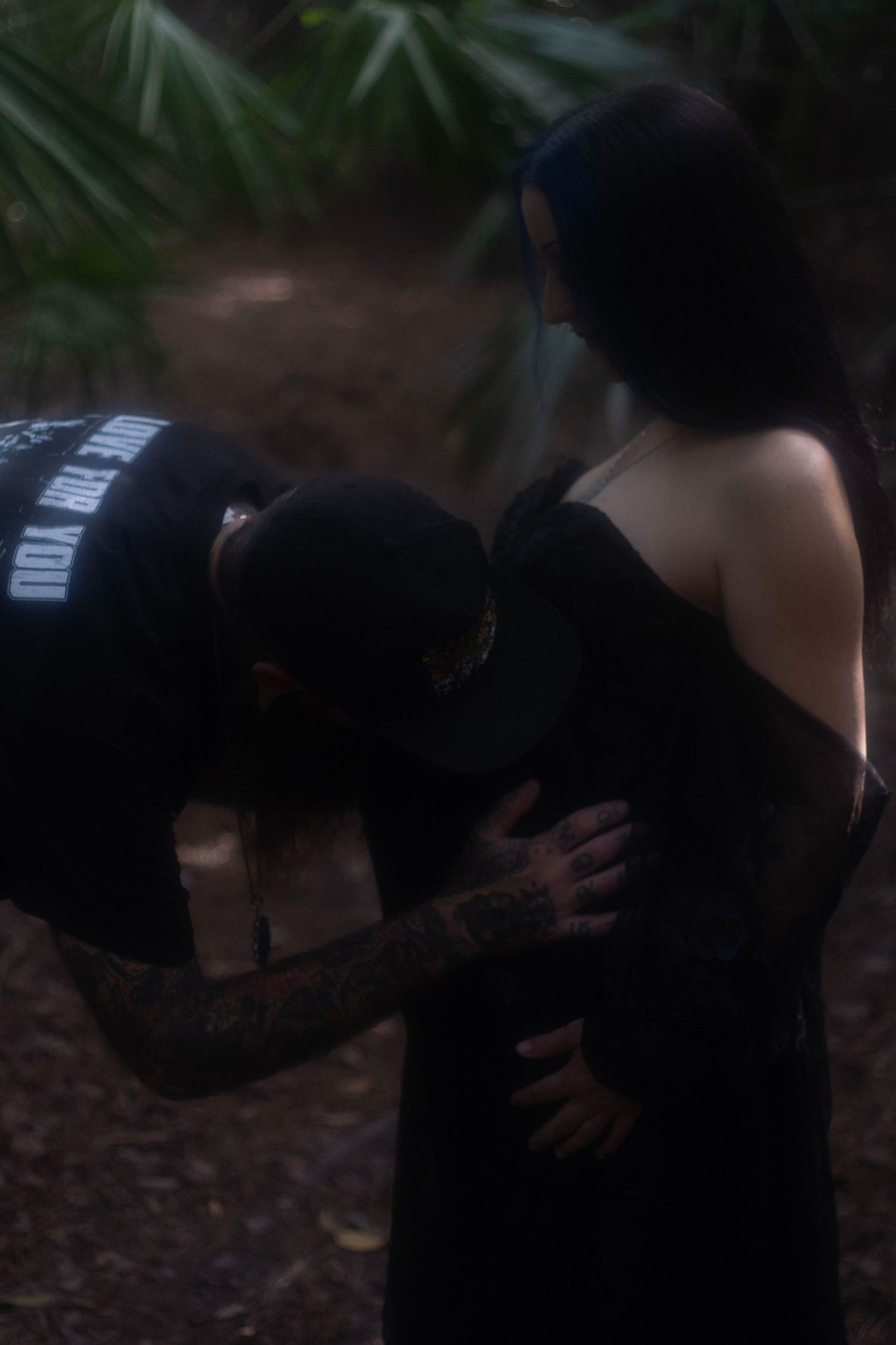 Tattooed man kissing pregnant woman's belly in a dark forest maternity photo session.