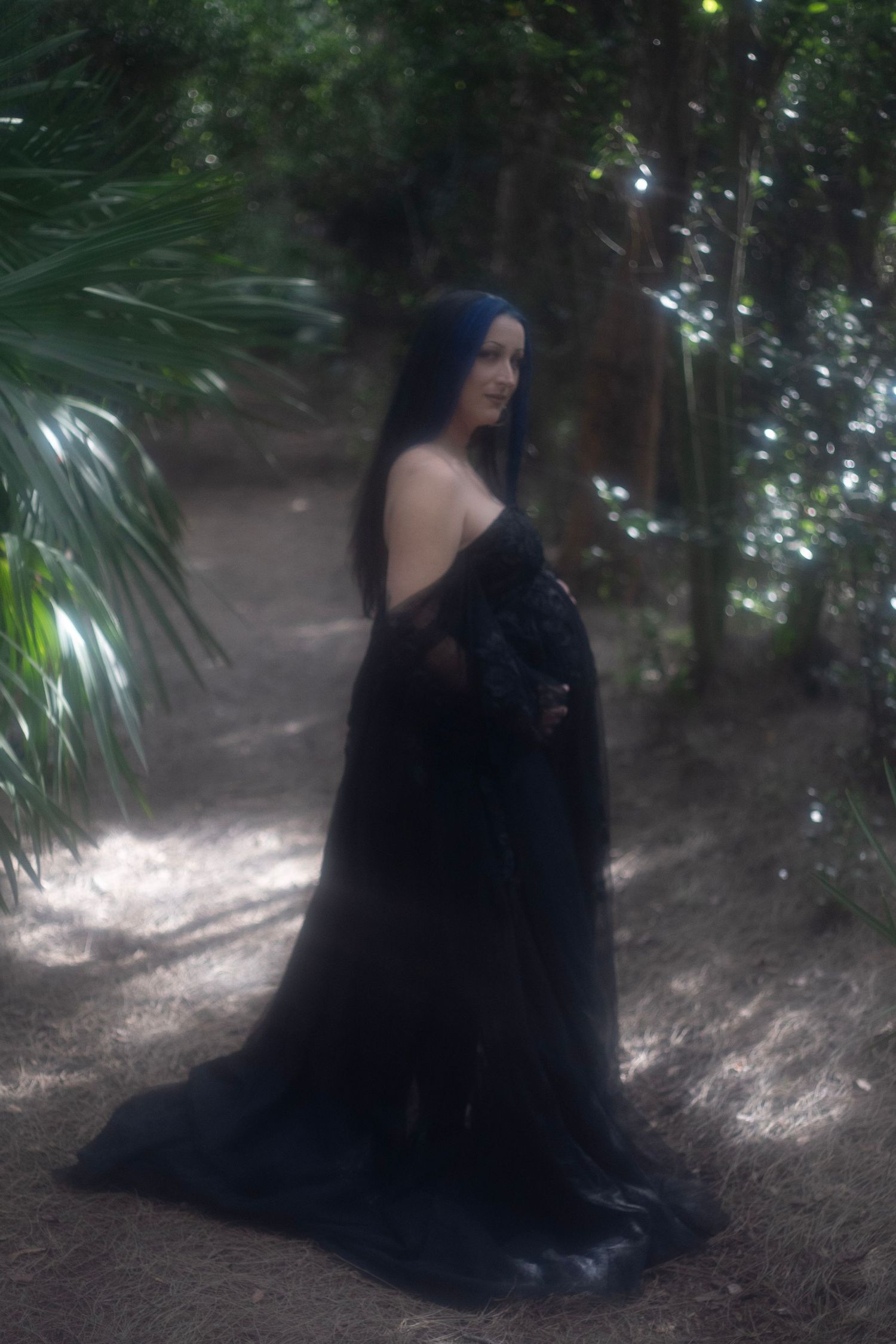 Pregnant woman in flowing black lace gown standing in a lush forest with soft dreamy lighting.