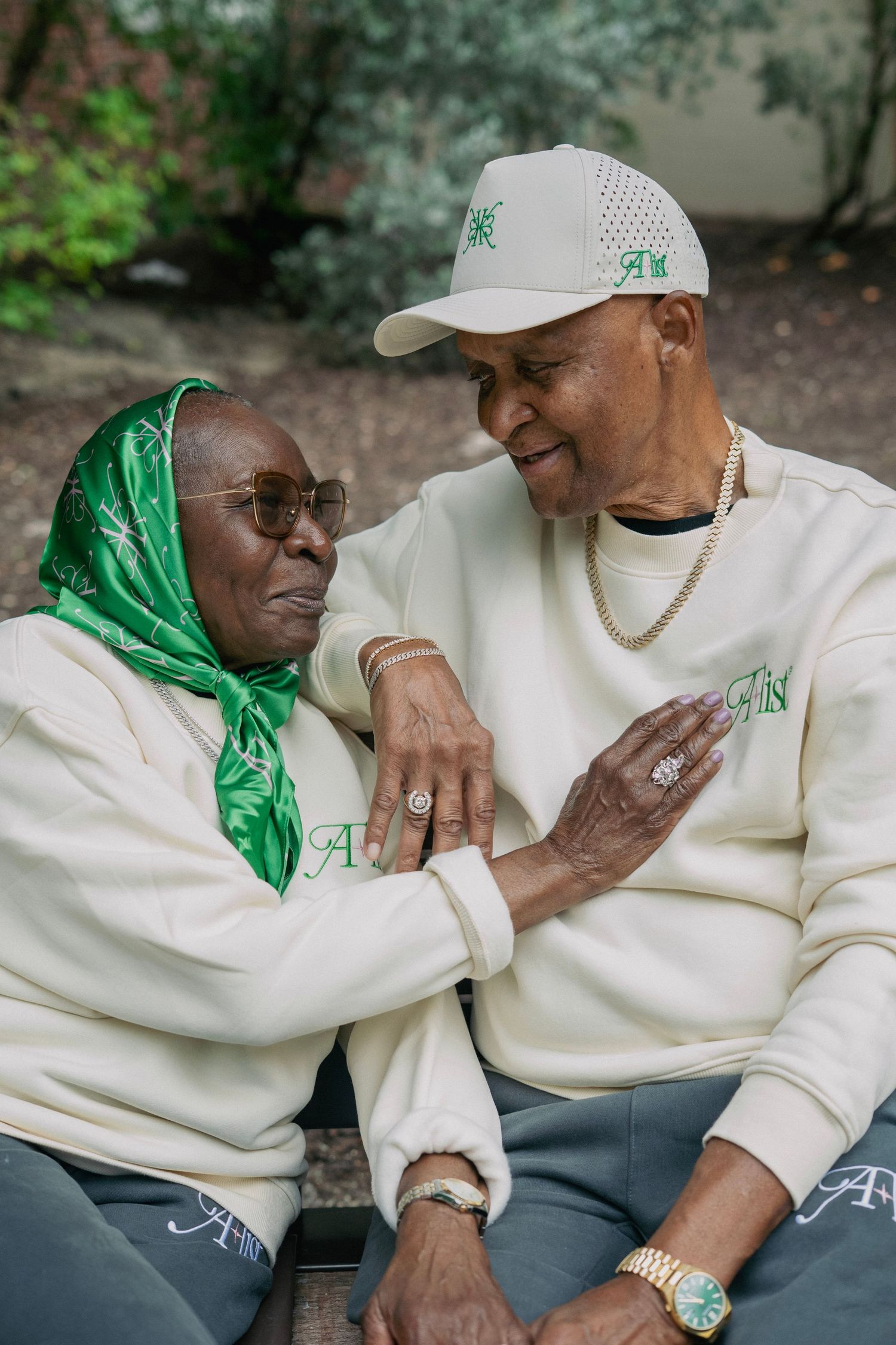 Two elderly Black people in matching cream sweatshirts smiling warmly at each other outdoors.