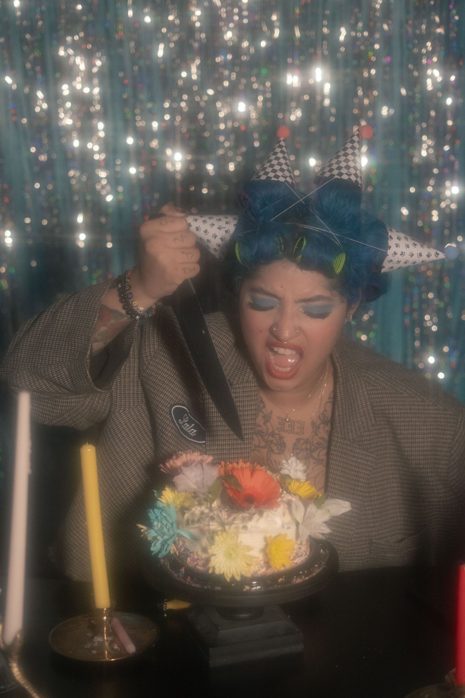 Woman with blue hair raises a knife over a floral cake with an intense expression and sparkly backdrop.