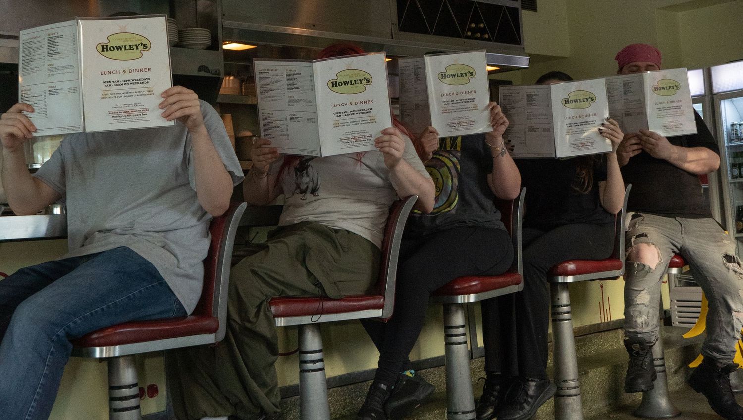 Four people sitting at a diner counter holding up Rowley's restaurant menus in front of their faces.