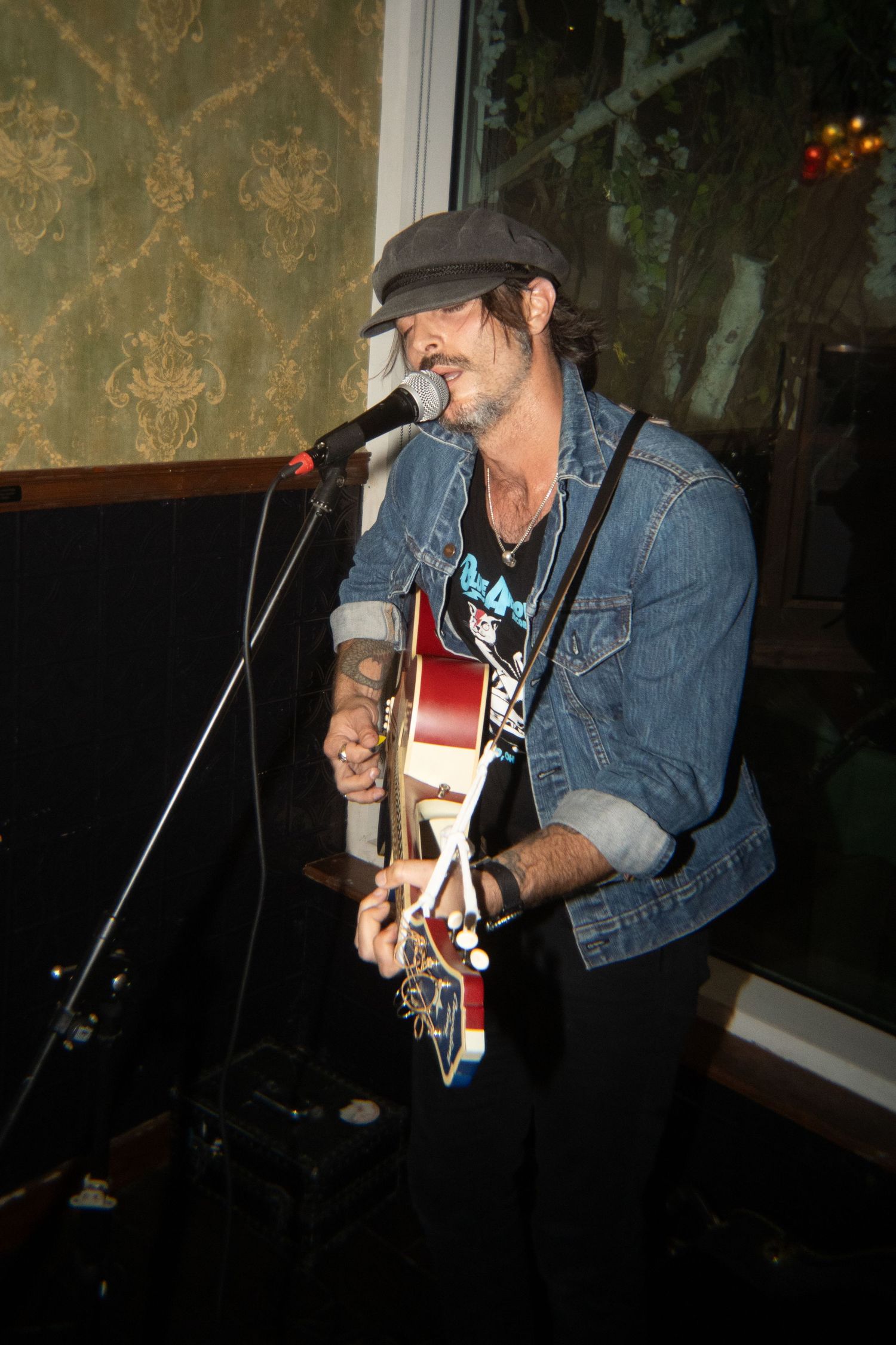 Male musician playing red guitar and singing into microphone, wearing denim jacket and gray cap at indoor venue.