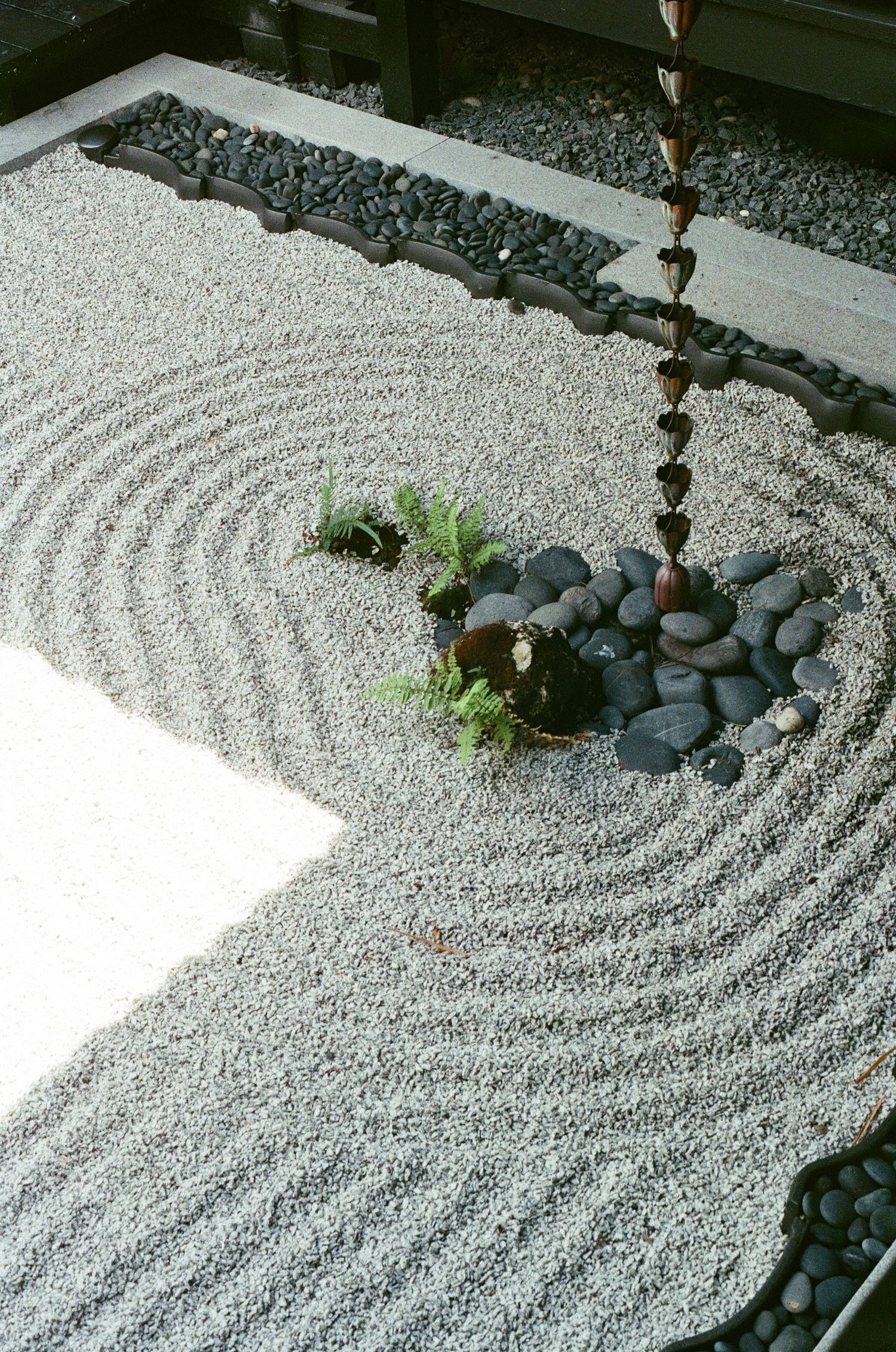Aerial view of a serene Japanese zen garden with raked gravel patterns, black stones, and a decorative chain water feature.