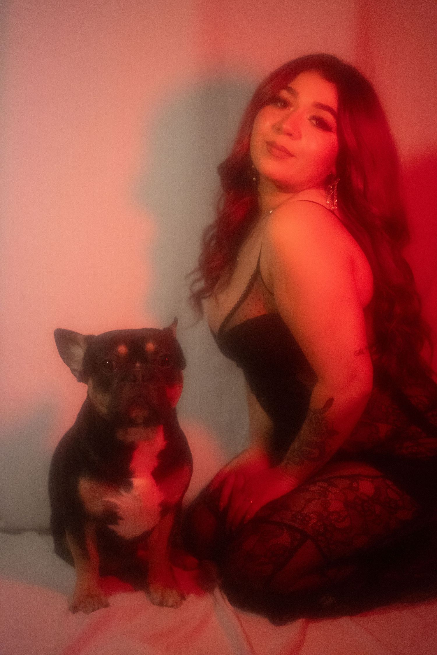 Woman with red hair posing beside a black French bulldog under red ambient light.