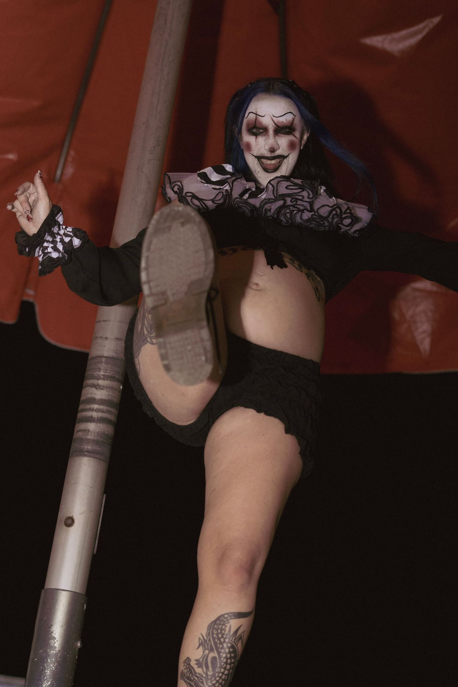 Pregnant woman in clown makeup and blue pigtails performs a high kick near a red tent at night.