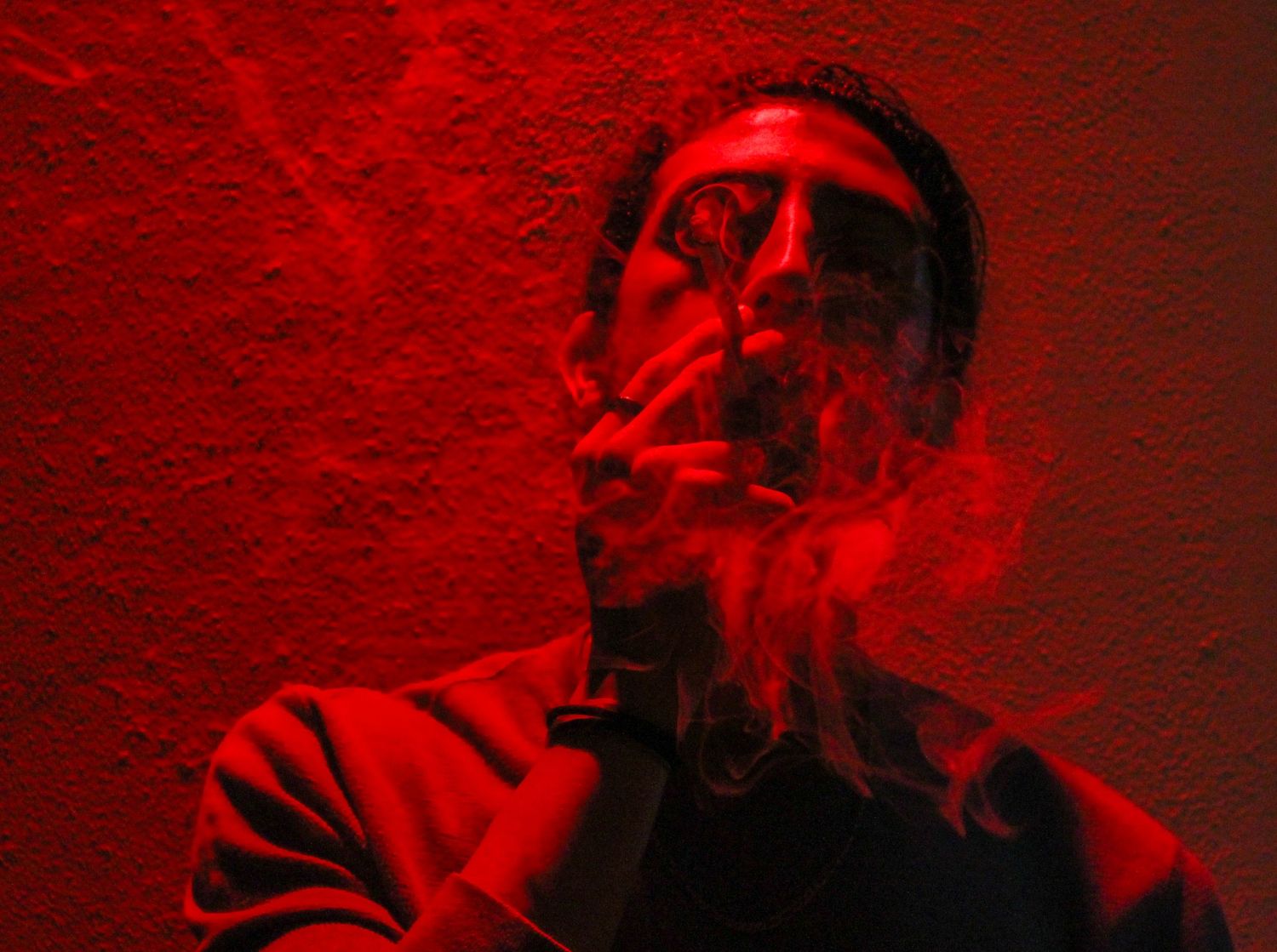 Man smoking a cigarette bathed in dramatic red light against textured wall creating moody atmospheric portrait.