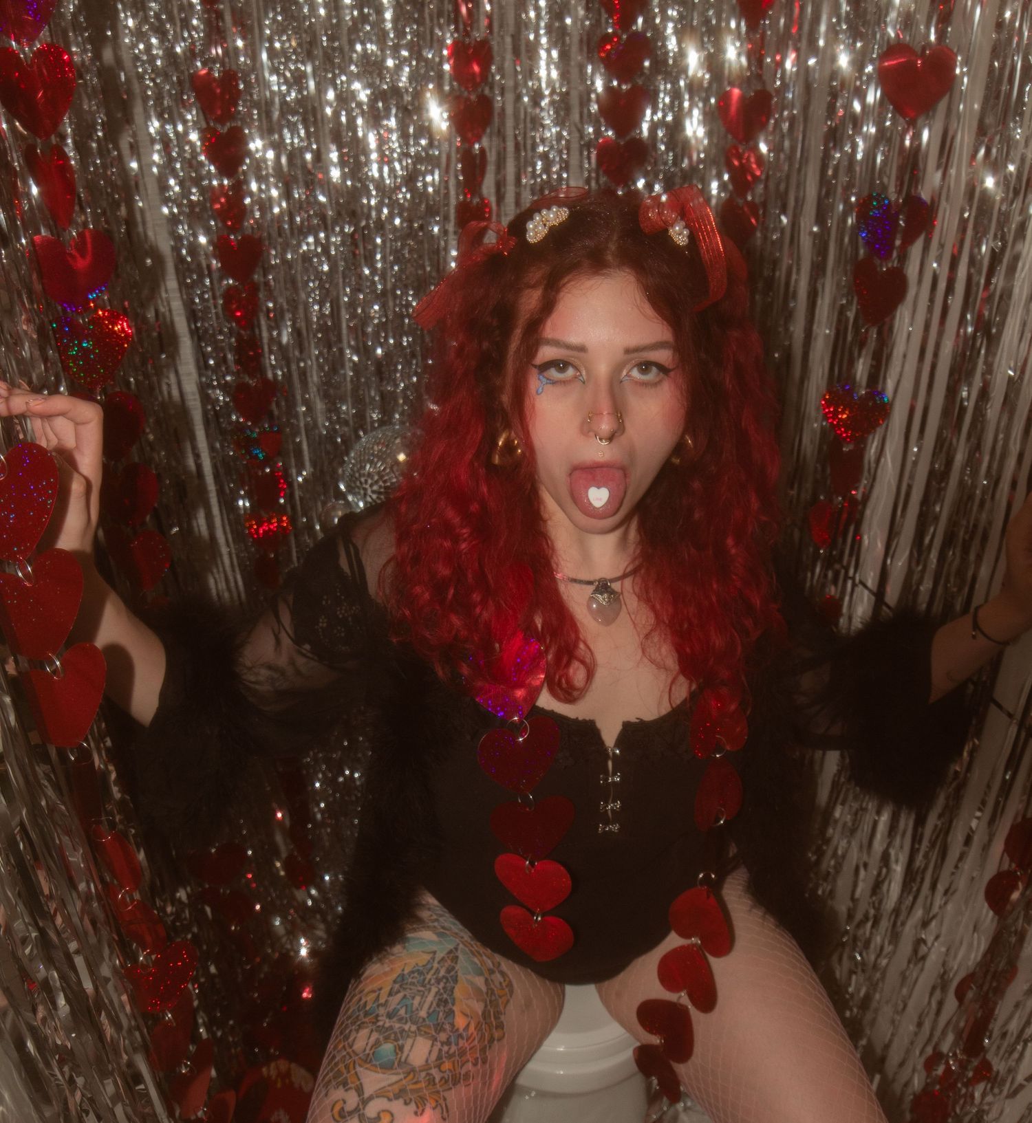 Red-haired woman in black top posing among silver tinsel and red heart decorations in festive Valentine-themed setting.