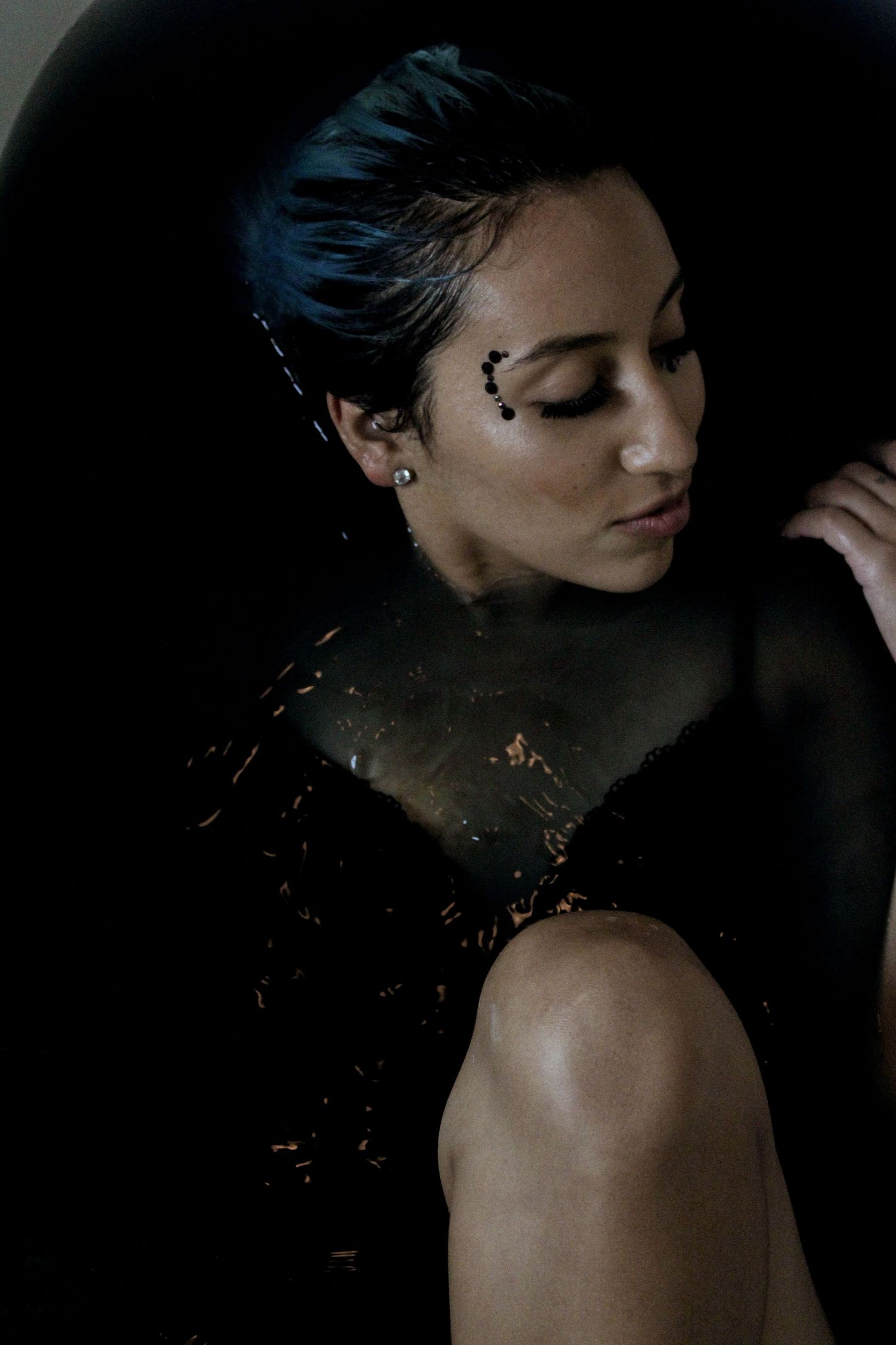 A woman with short dark hair and black face gems sits in a dark bathtub, golden particles shimmering around her.