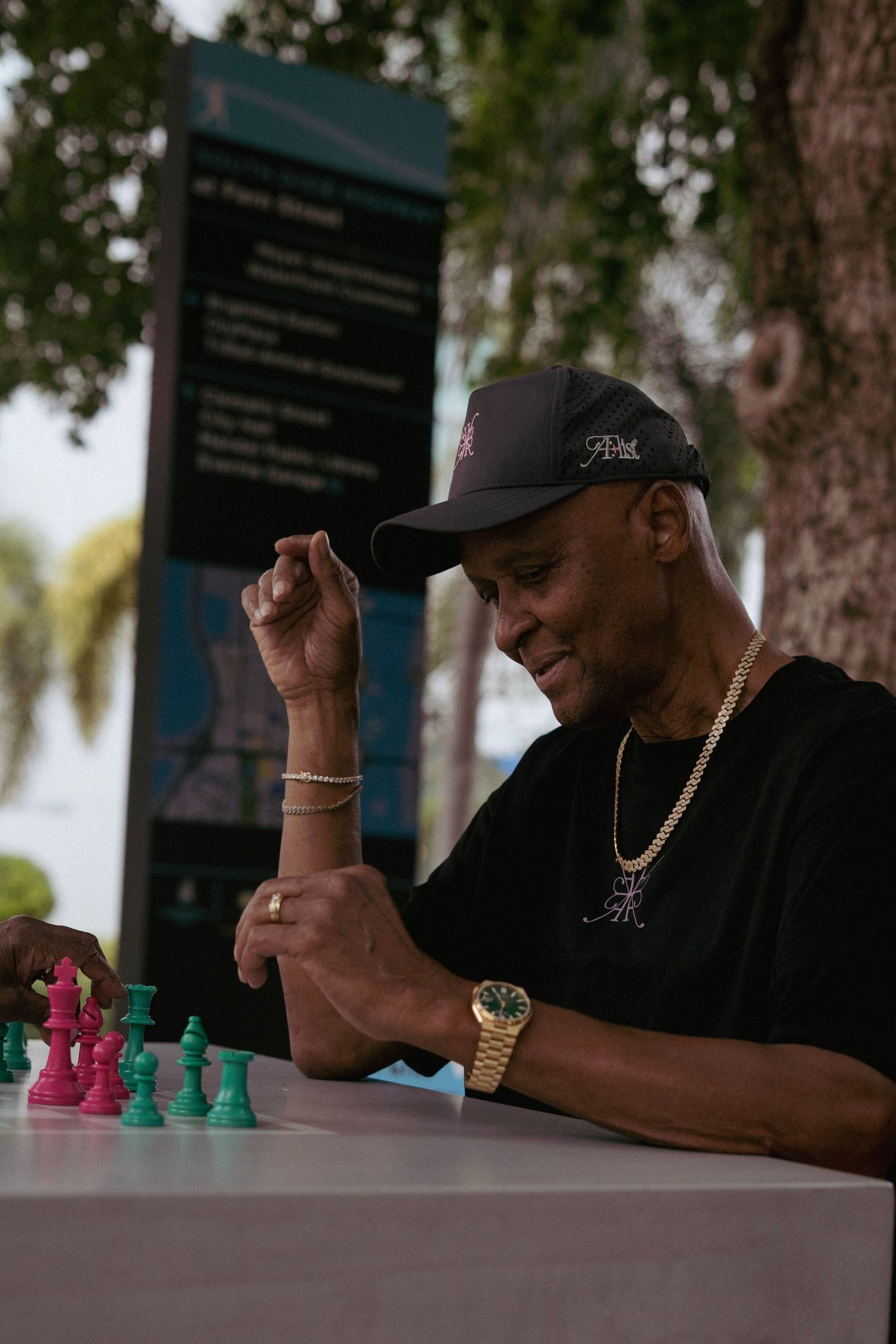 Black man in dark cap and gold chain playing chess with pink and green pieces outdoors.