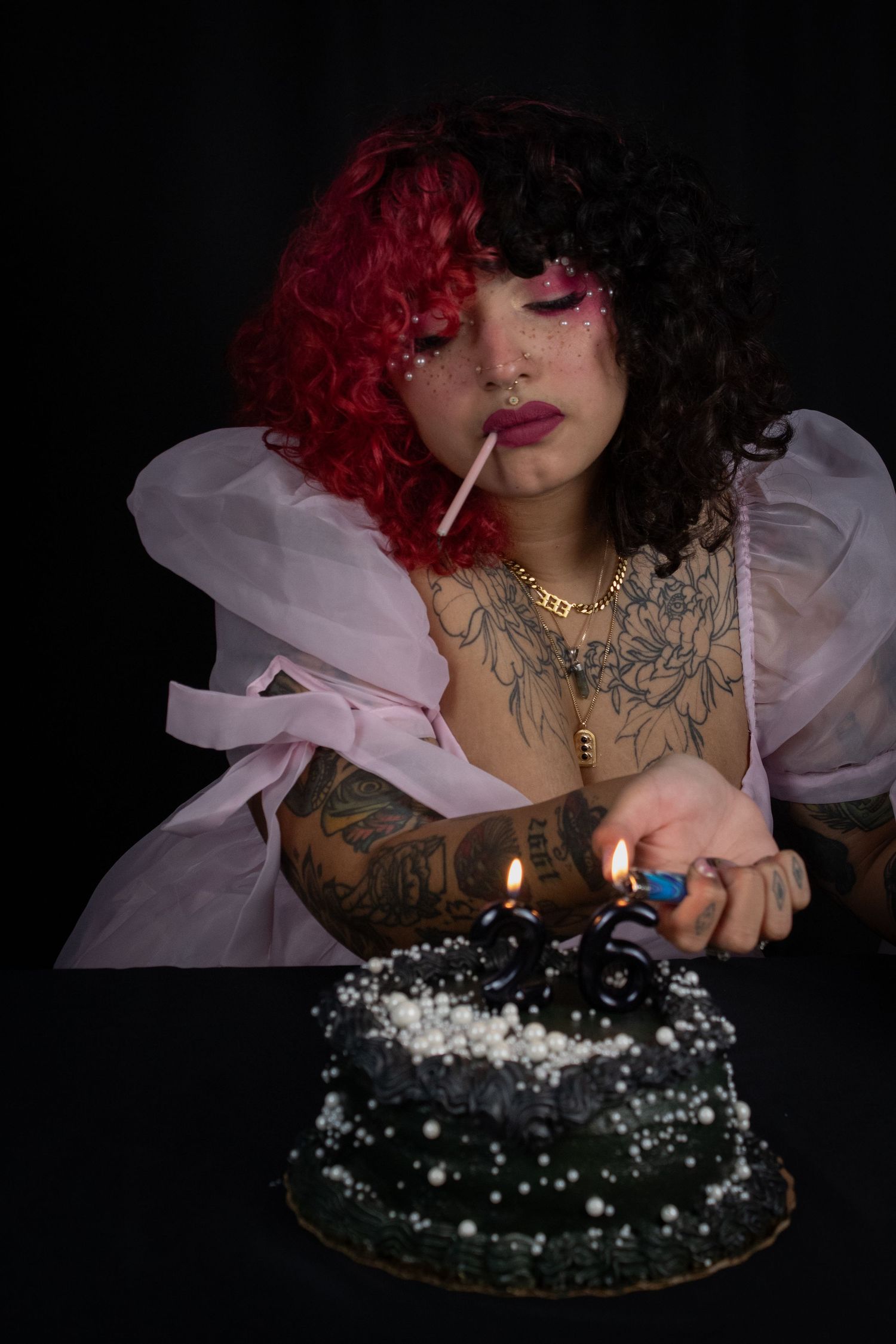 Tattooed woman with red and black curly hair lighting candles on a dark birthday cake while holding a lollipop.