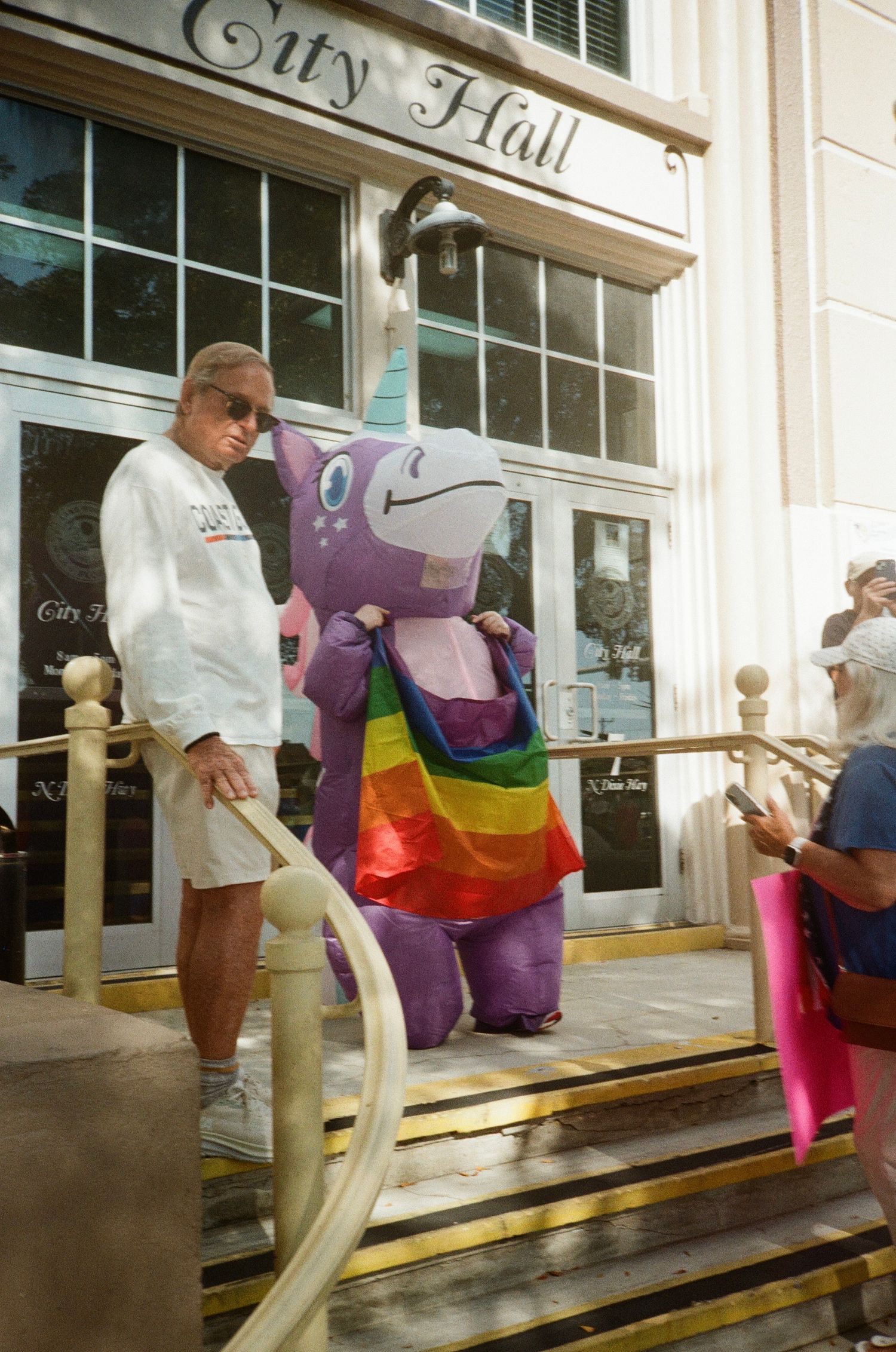 A person in a colorful inflatable rainbow unicorn costume stands outside a city hall building.