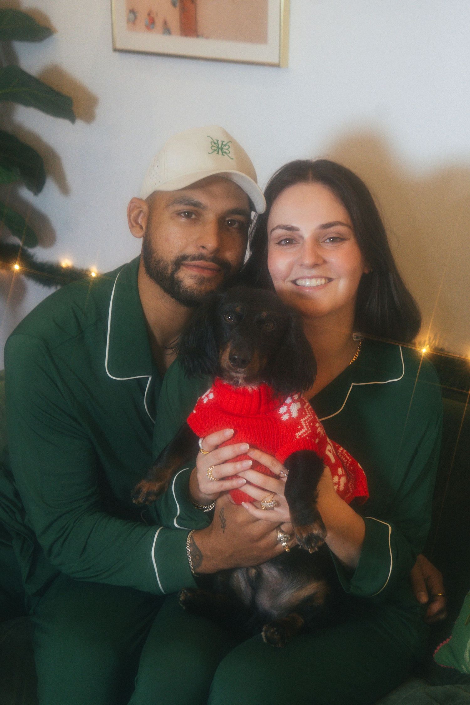 Couple in matching green Christmas pajamas holding a dachshund in a red holiday sweater.