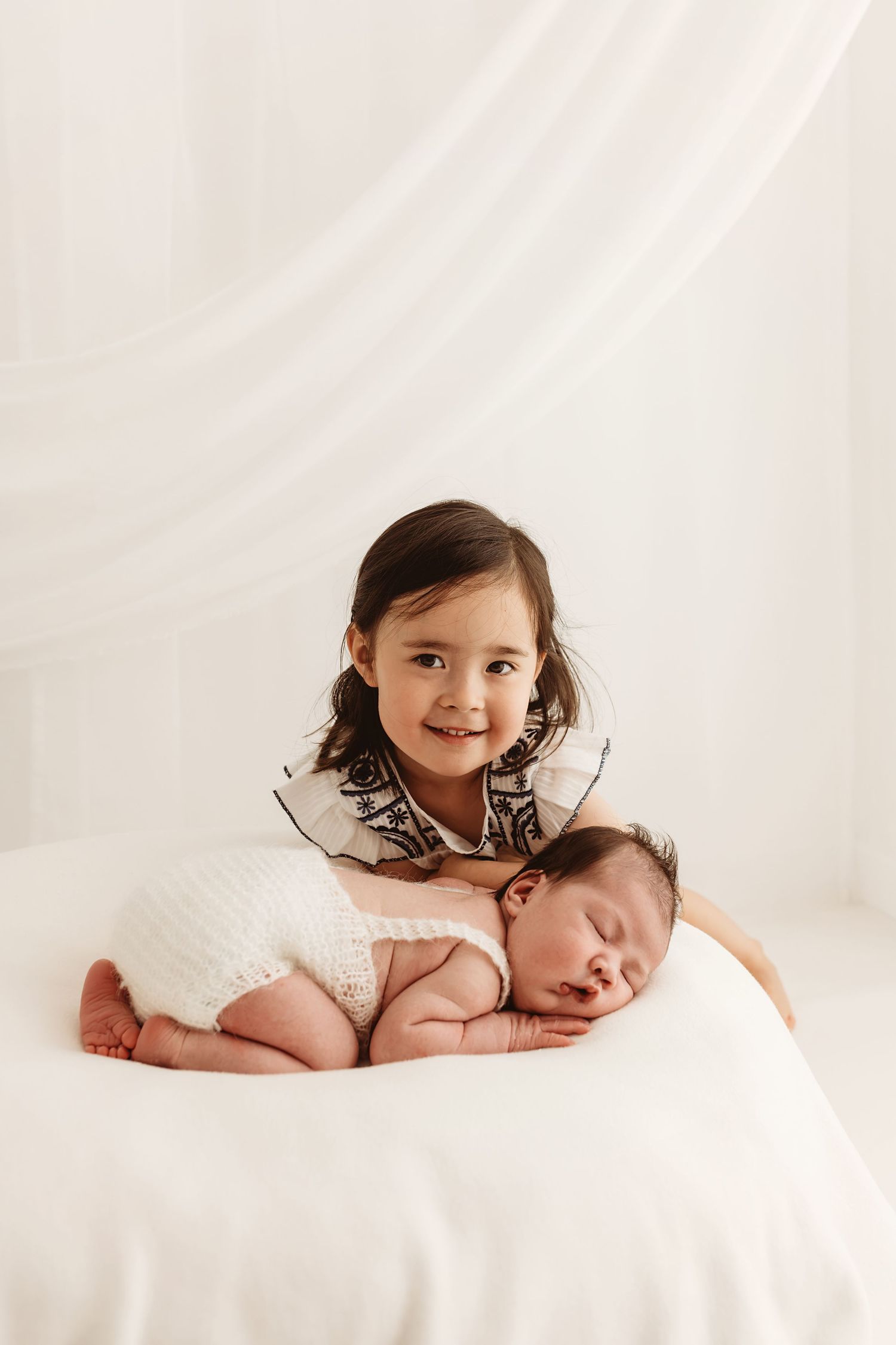 A smiling toddler girl in a patterned dress poses with her sleeping newborn sibling wrapped in white on a soft white surface.
