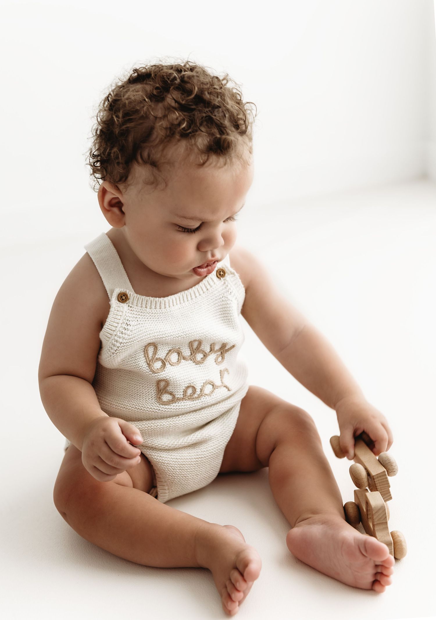 Curly-haired baby in a cream knit Baby Bear romper sits playing with wooden toy blocks during a Sitter photography session in Corby