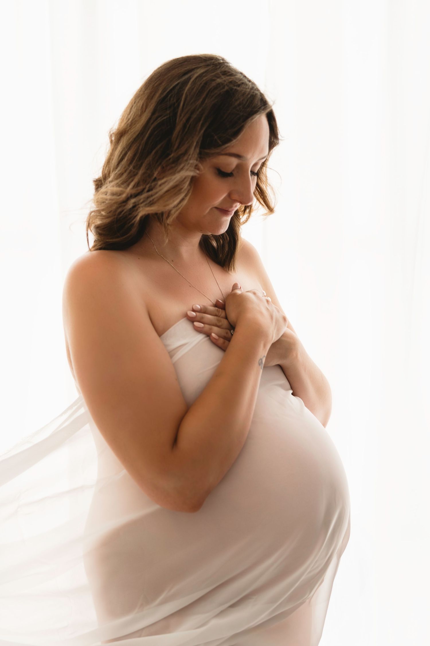 Ethereal maternity portrait of woman draped in white fabric against light background.