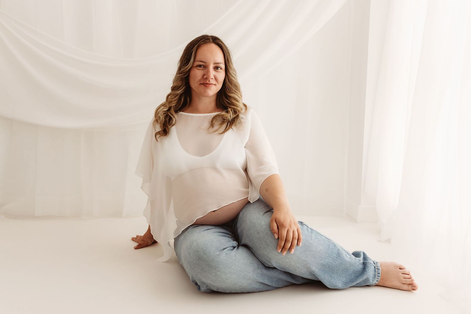 Pregnant woman in white sheer top and jeans sitting cross-legged on white studio floor, smiling softly.