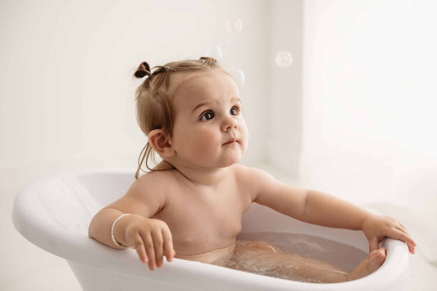 Cute baby girl sitting in a small white bathtub with bubbles, enjoying bath time in a bright bathroom.
