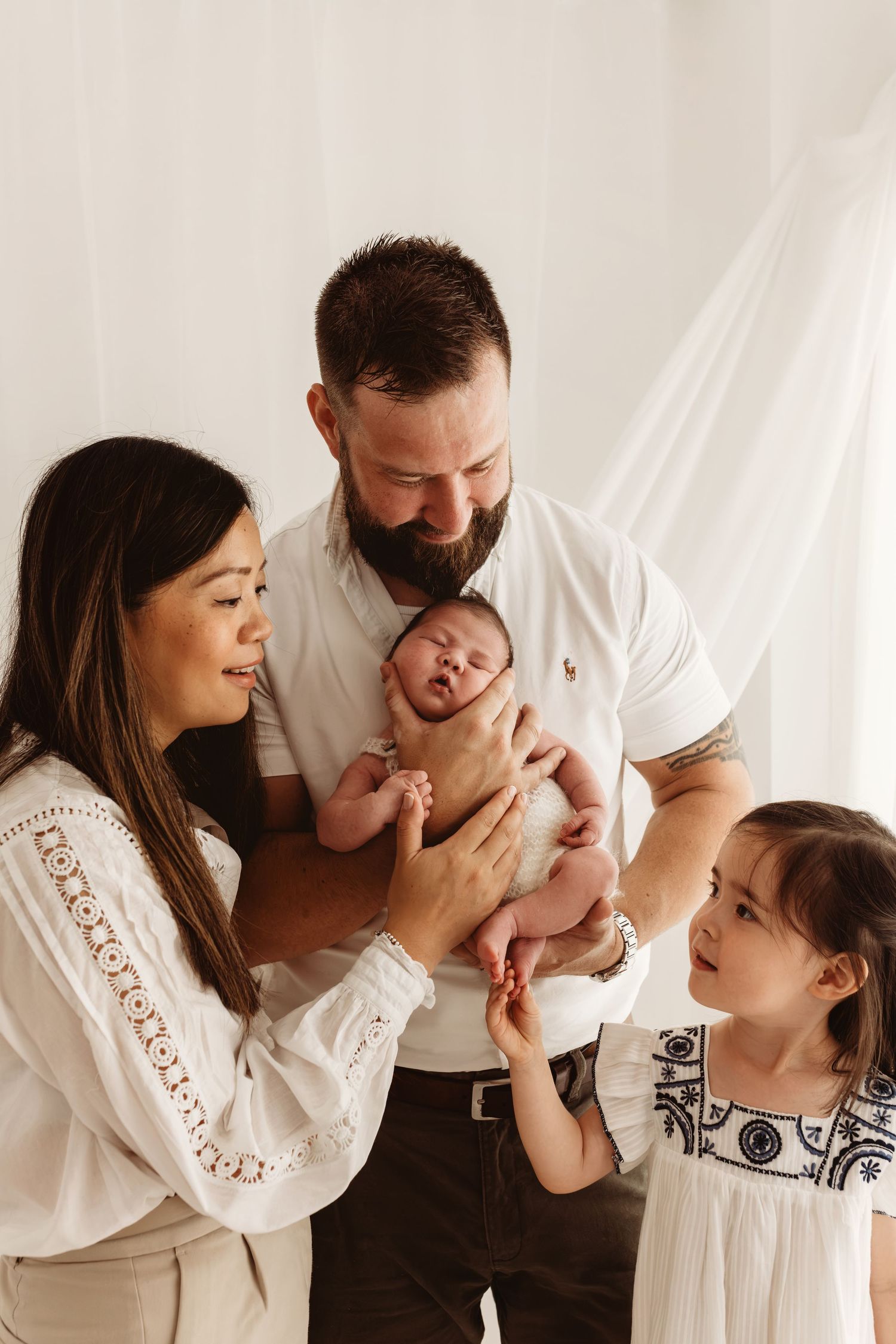 Family of four with parents and young girl admiring newborn baby in bright white studio setting.