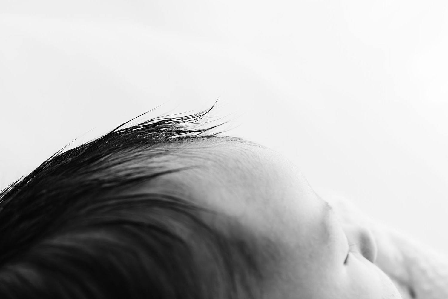Close-up black and white photo of a newborn baby's head with fine dark hair against a white background.
