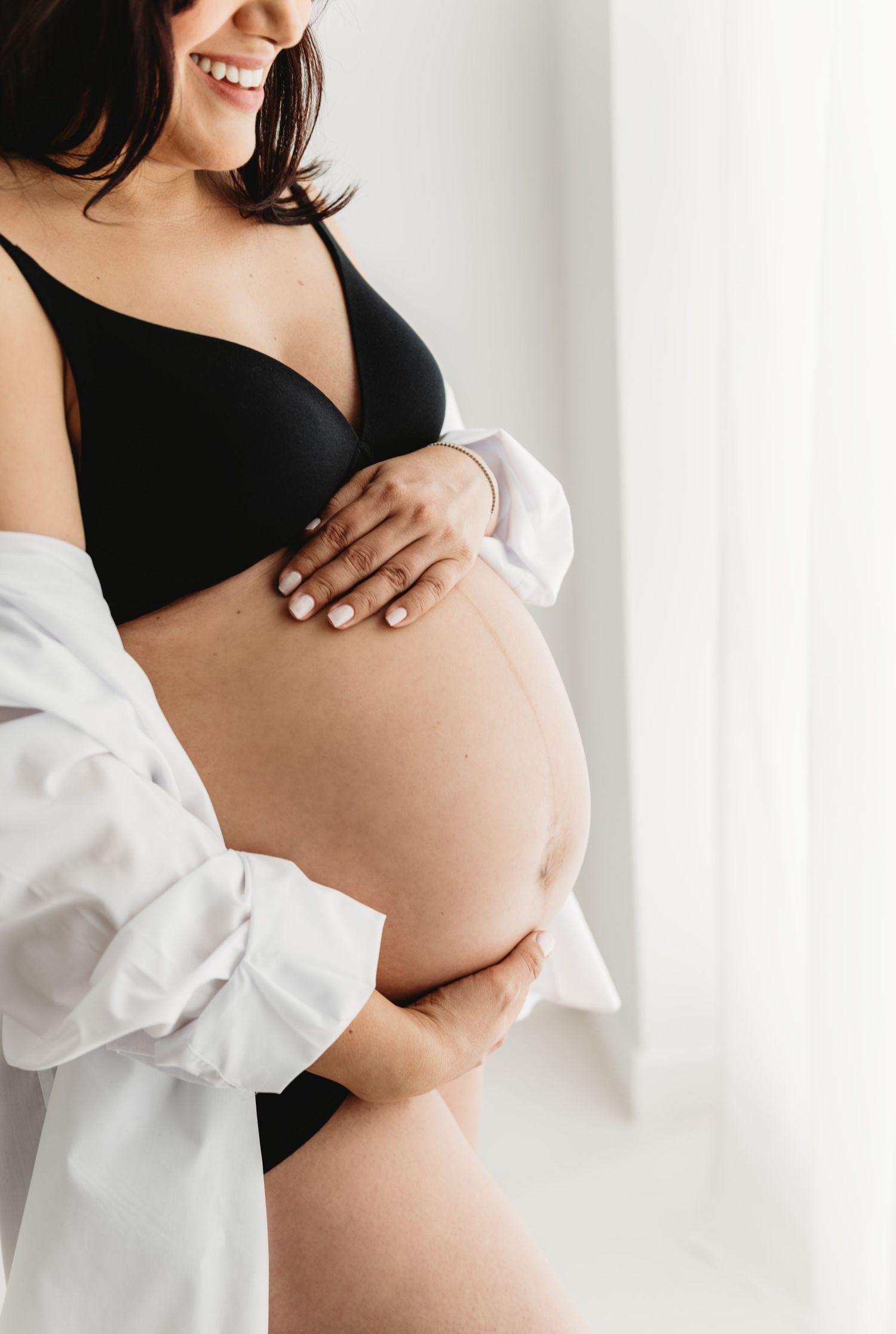 Smiling pregnant woman in black bra and underwear cradling her baby bump, wearing an open white shirt.