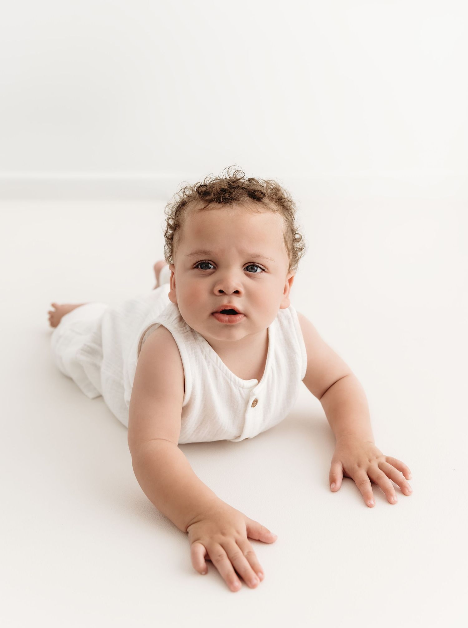 Baby wearing white sleeveless outfit lying on white surface looking at camera with curious expression.