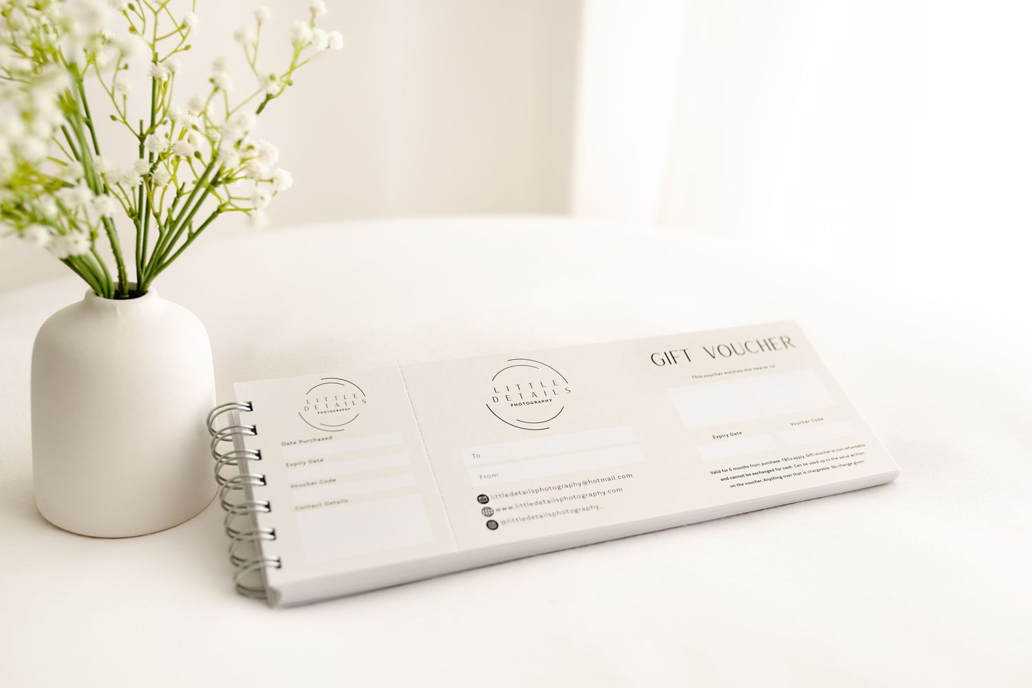 A white ceramic vase with delicate flowers sits next to a spiral-bound notebook on a clean white surface.