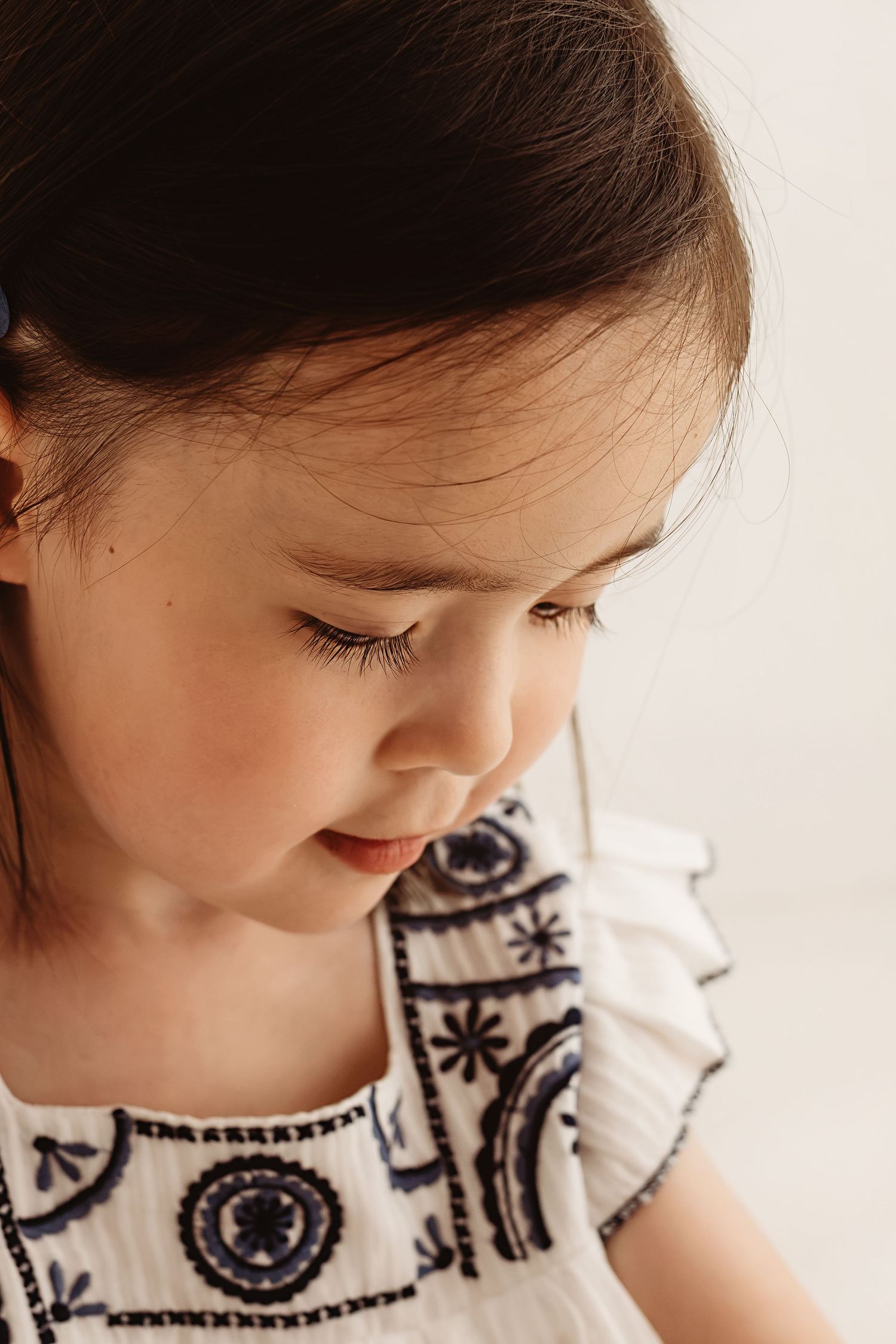 Adorable young girl with dark hair looking down, wearing white embroidered dress in soft light.