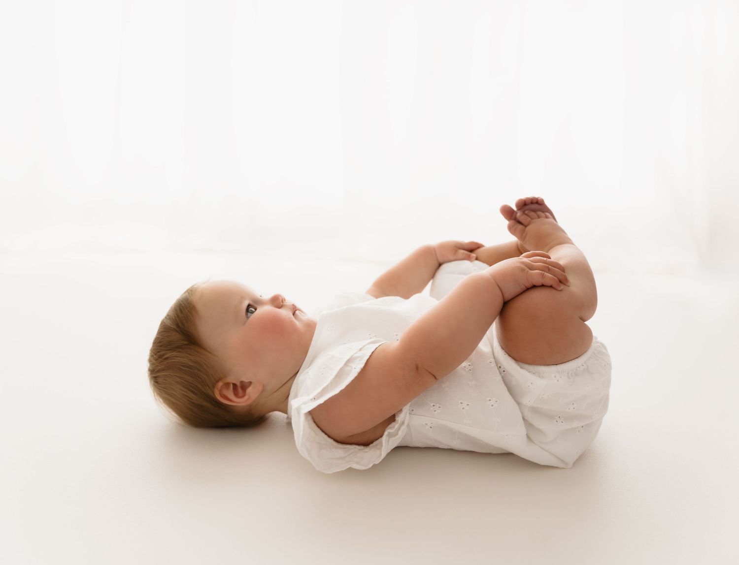 Cute baby in white onesie lying on back, holding feet up, on a bright white background.