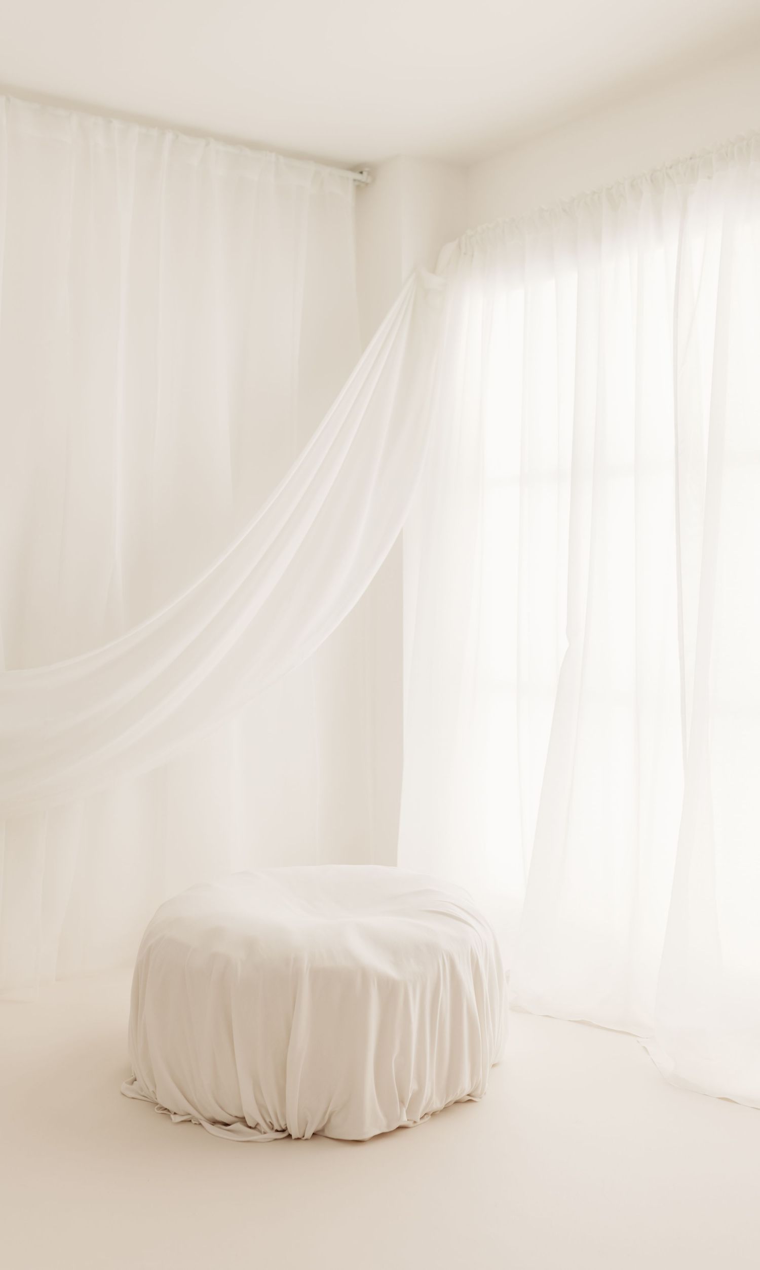 Ethereal white draped fabric covers a round ottoman in a minimalist room with flowing sheer curtains and soft natural light.