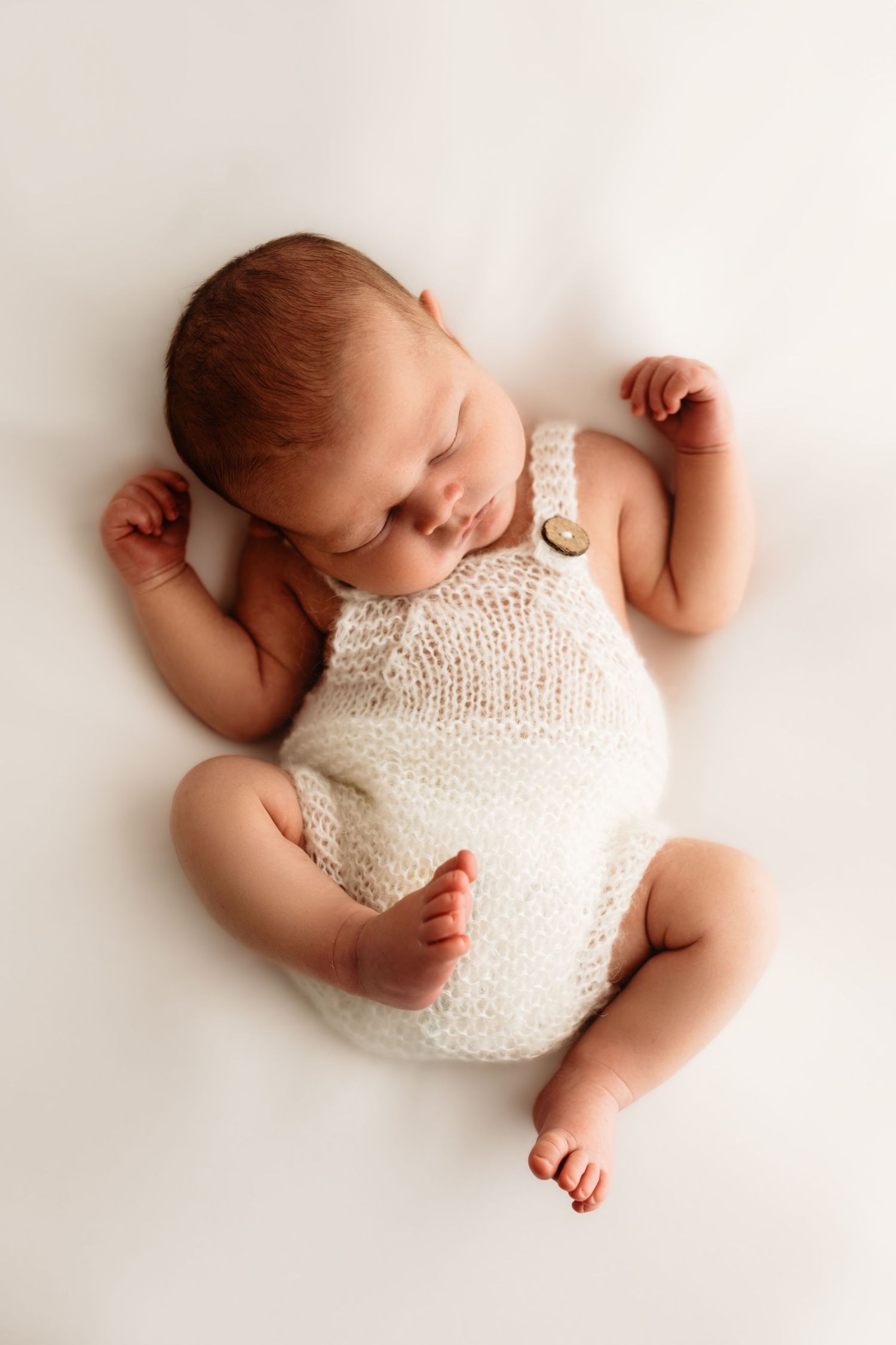 Sleeping newborn baby wearing white knit romper lying on white backdrop in professional studio portraits.