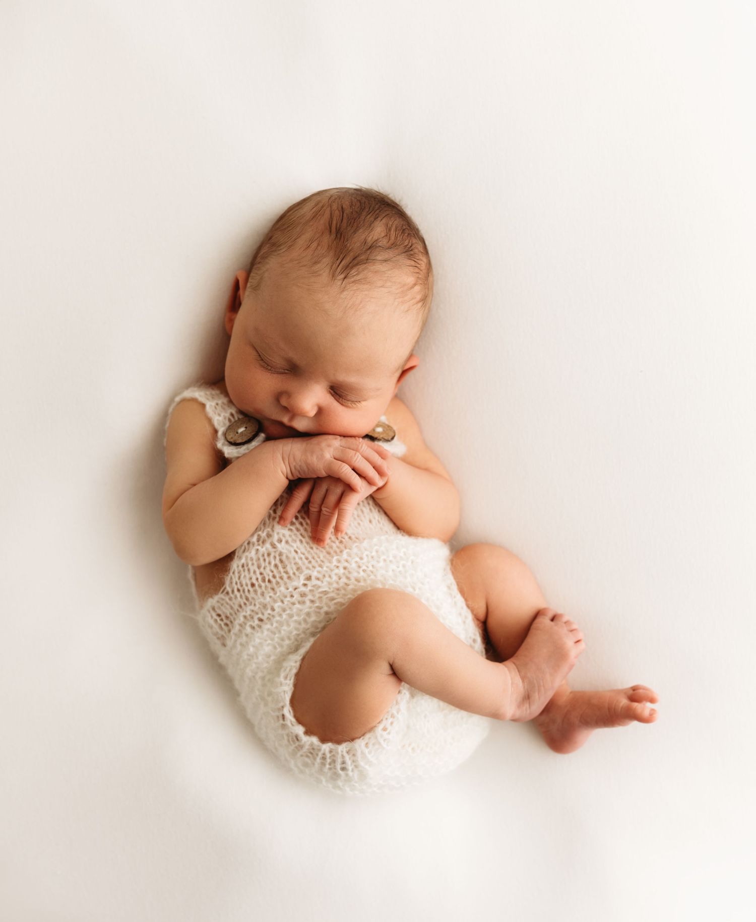 Newborn baby sleeping peacefully in white knit romper, curled up on soft white background in professional photo.
