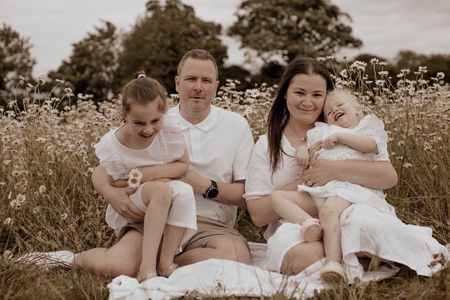 Family of four sitting in a wildflower meadow, parents holding young children, wearing white outfits in a serene outdoor setting.