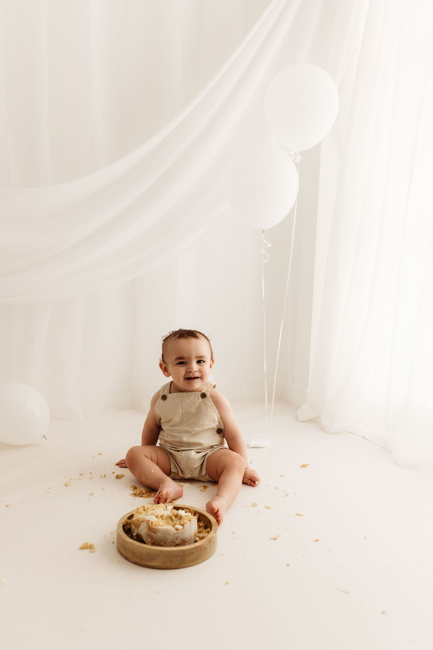 Baby in beige romper sits on white floor next to wooden cake plate during minimalist cake smash photo session.