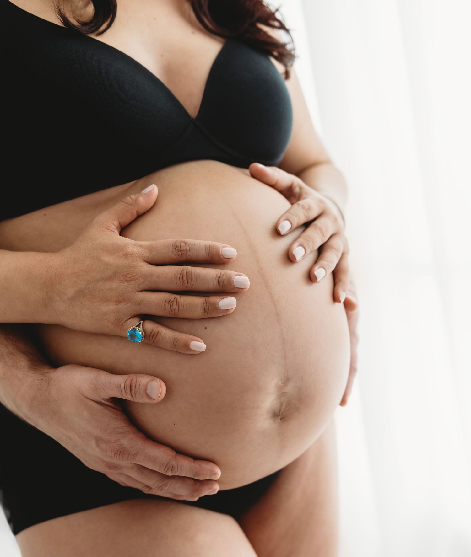 Pregnant woman in black lingerie cradling baby bump with partner's hands, maternity photography concept.
