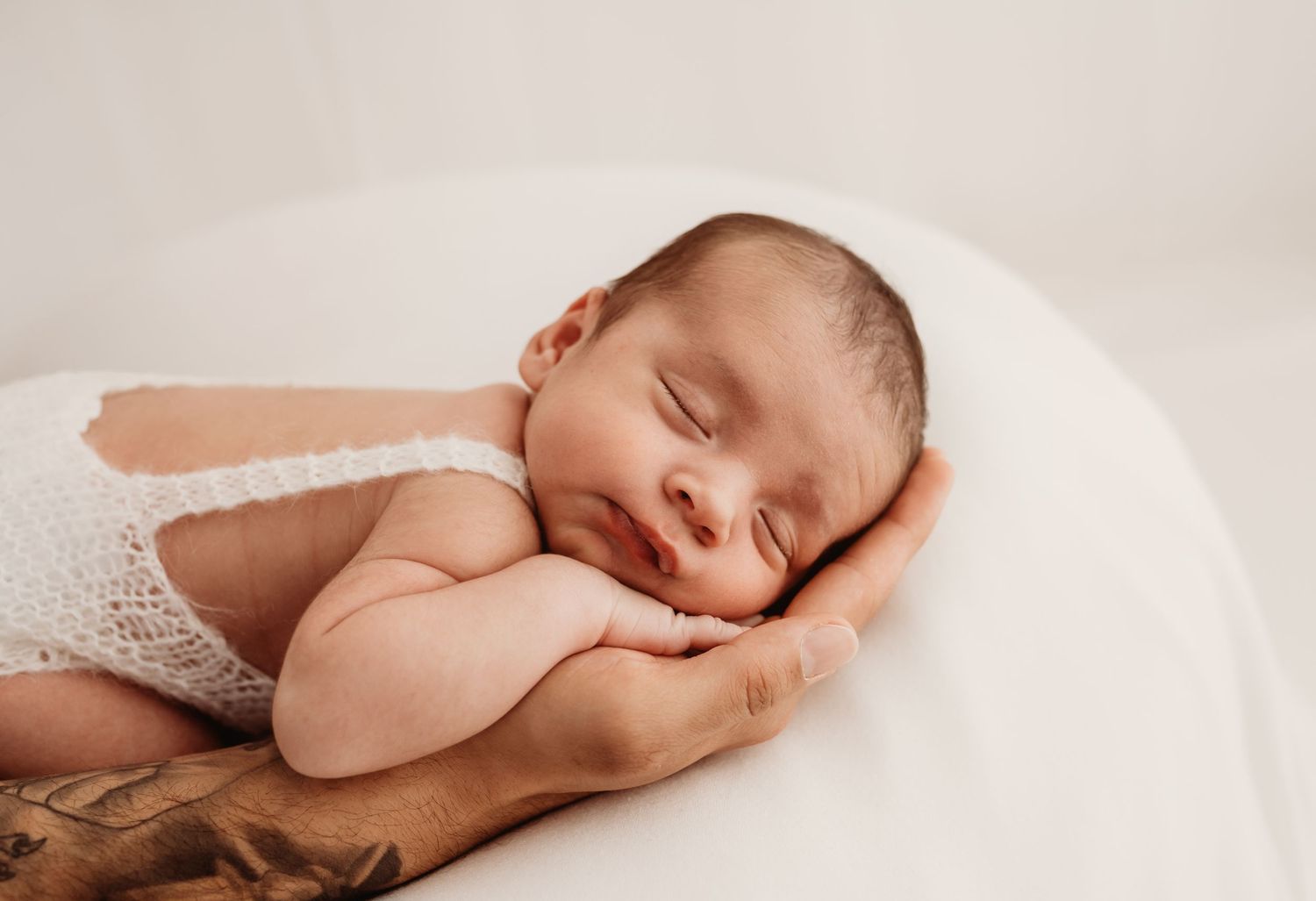 Peaceful newborn baby sleeping on adult hands wearing white lace romper against soft white background.