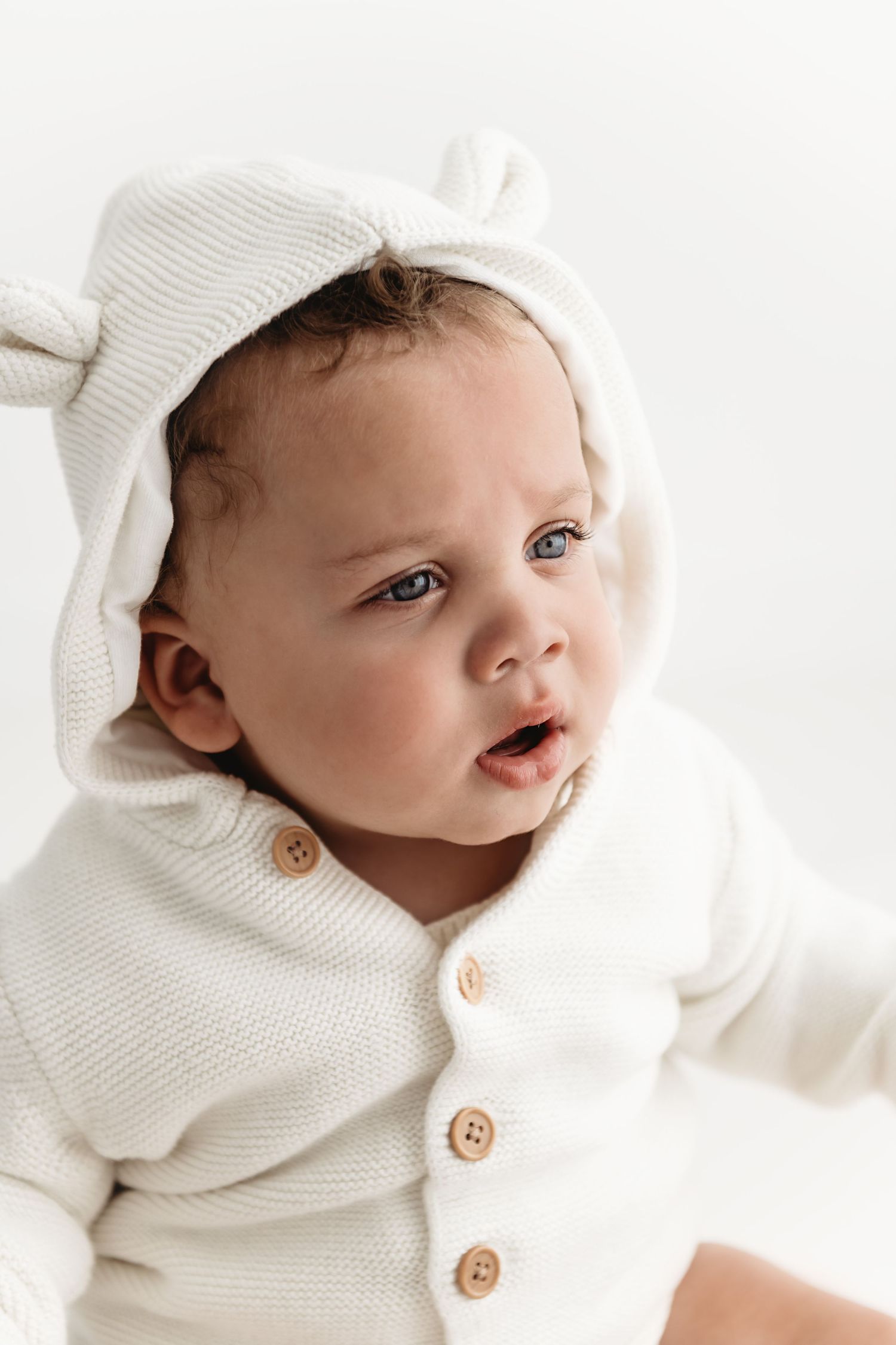 A small baby wearing a cozy white knit sweater with wooden buttons looks to the side with bright eyes.