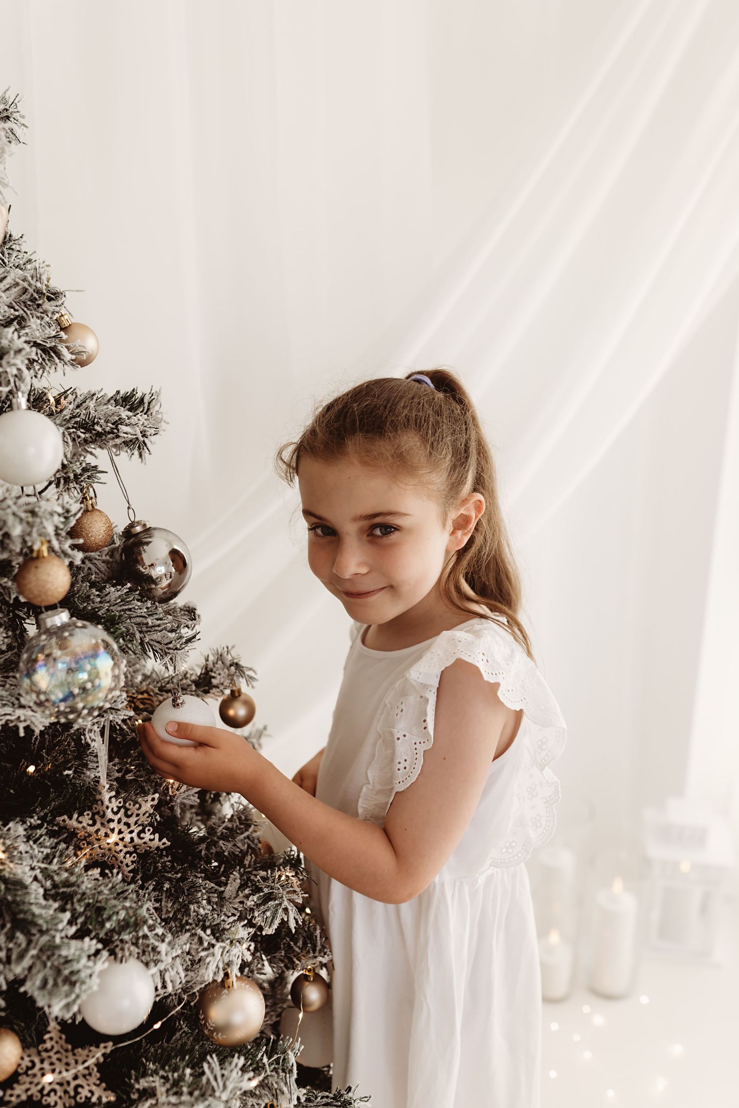 Young girl in white dress decorating a Christmas tree with gold and silver ornaments in a bright white room.