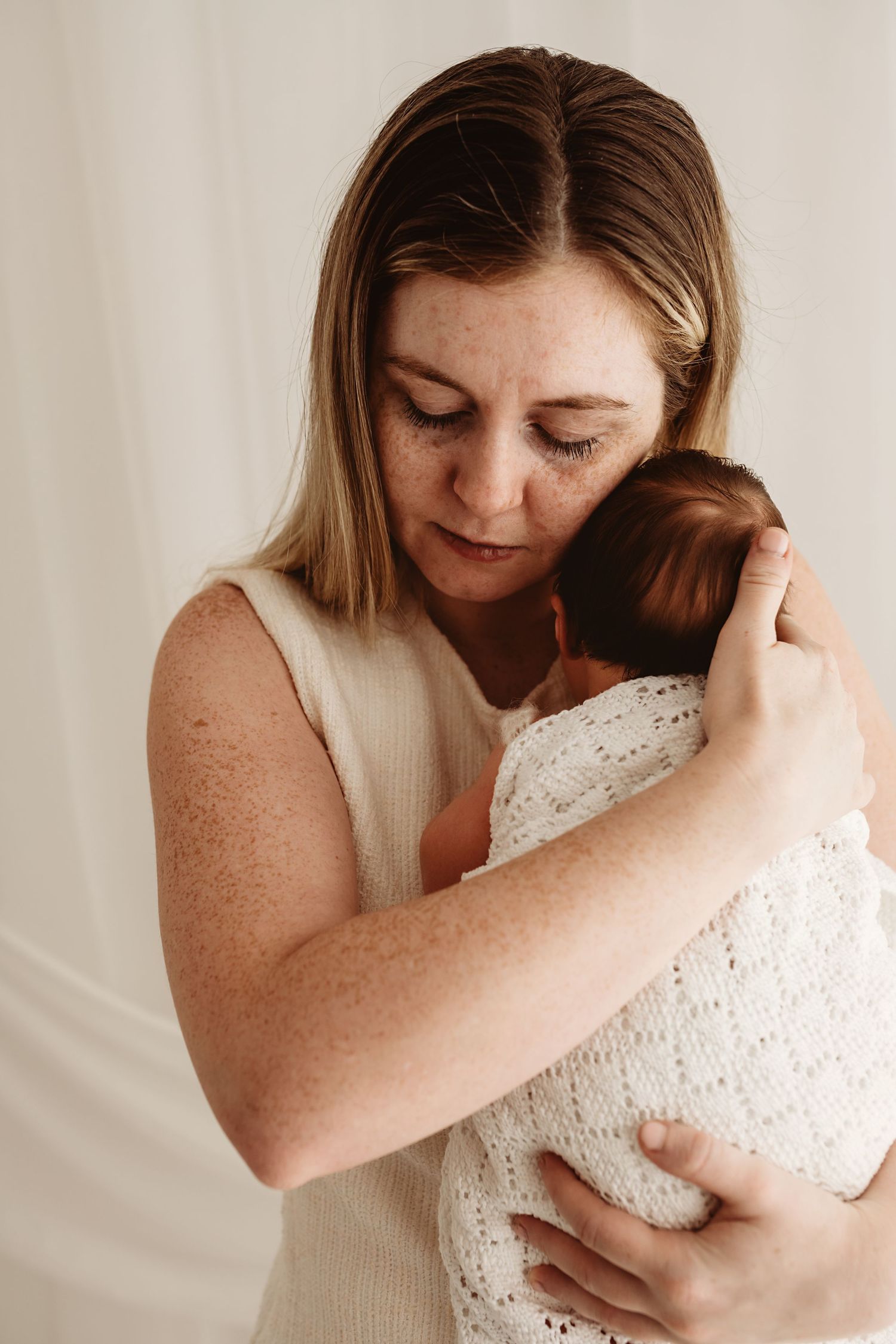 A freckled mother with blonde hair tenderly holds her swaddled newborn baby close to her chest in a soft, neutral setting.