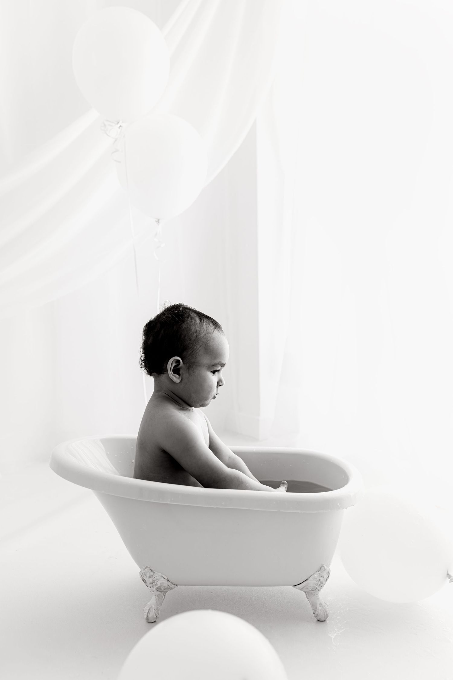 Black and white photo of a toddler sitting in a vintage clawfoot bathtub surrounded by balloons during a cake smash photography session