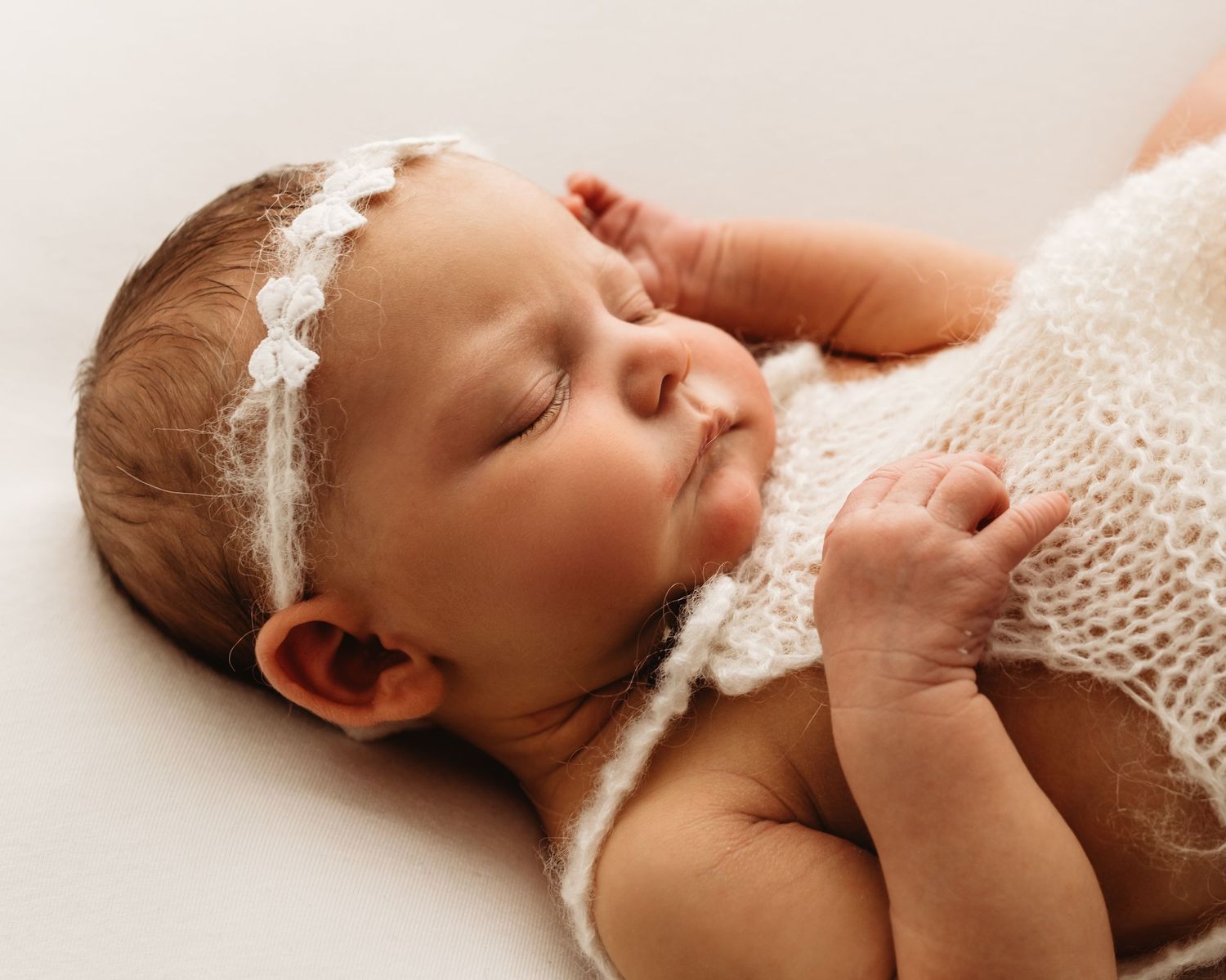 Peaceful newborn baby sleeping wrapped in white knit blanket with lace headband on white background.